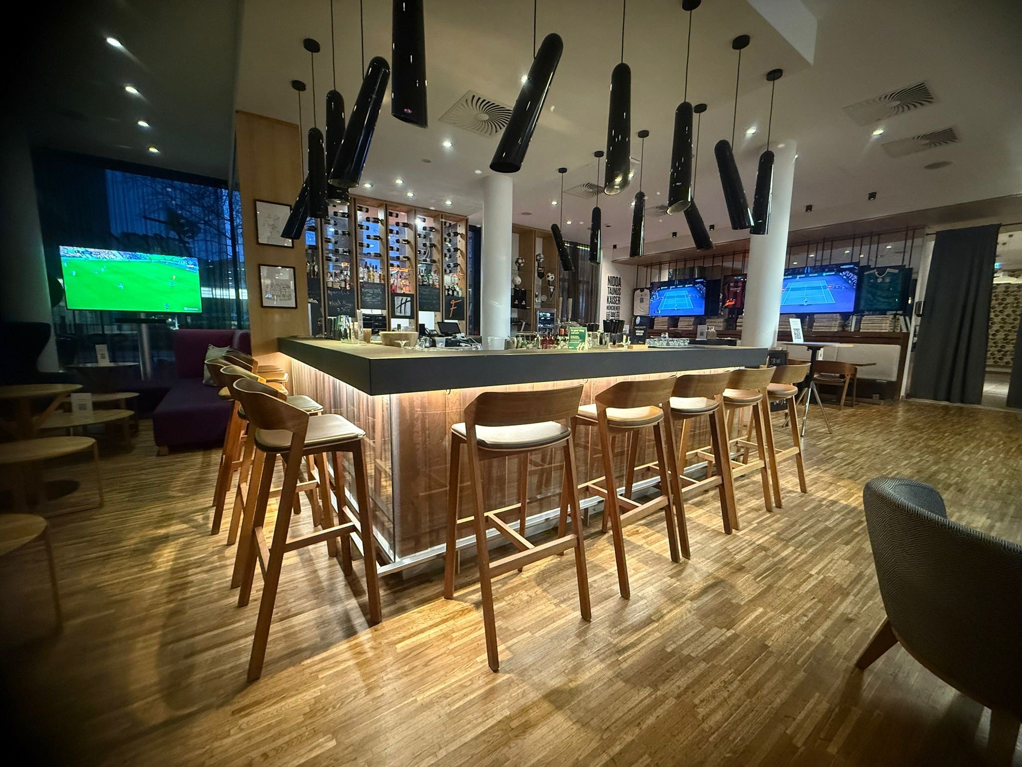 A modern bar with a high counter and stools, multiple TVs showing sports, and a wood floor in a well-lit lounge area.