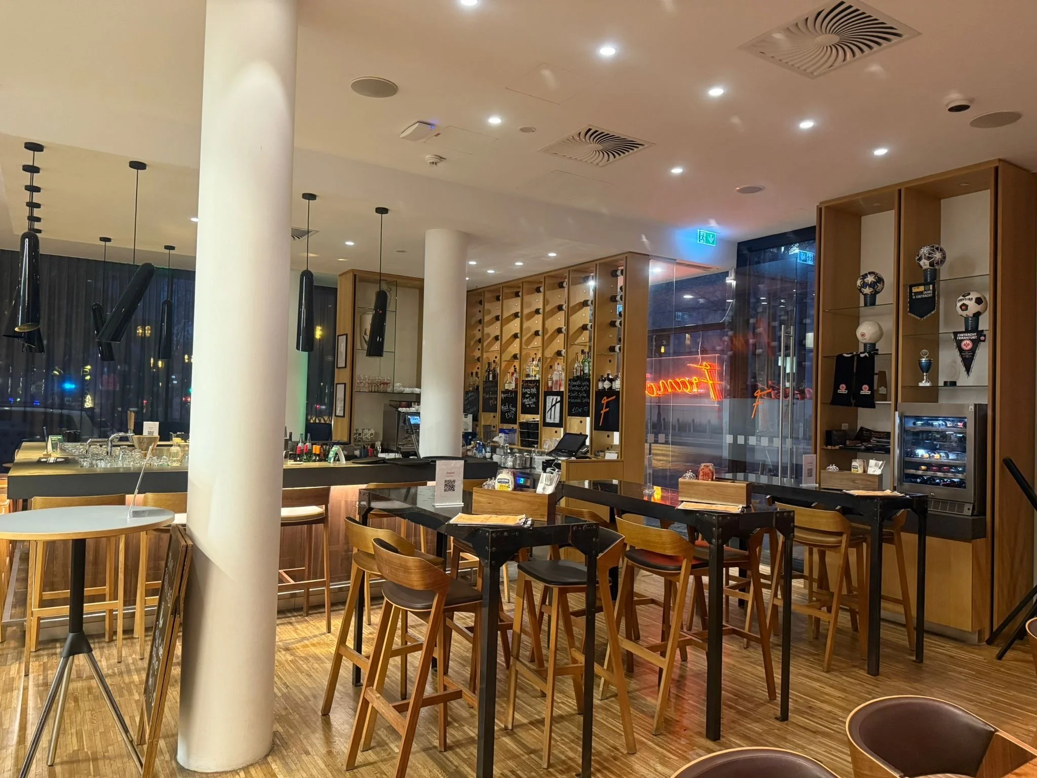 Interior of a modern bar or restaurant with wooden furniture, high stools, and a well-stocked bar area. There are some decorative items and a neon sign in the window.