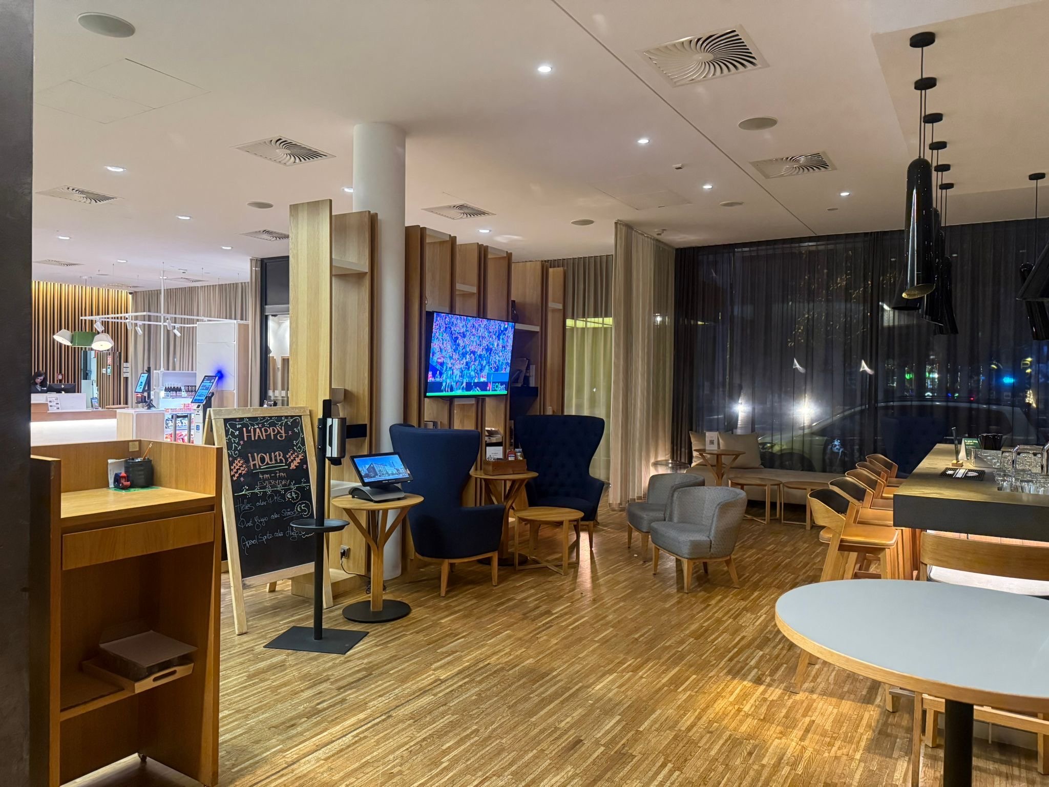 Interior of a modern café or lounge with various seating options, a television, a chalkboard sign, and large windows with dark curtains, illuminated by ceiling and pendant lights.