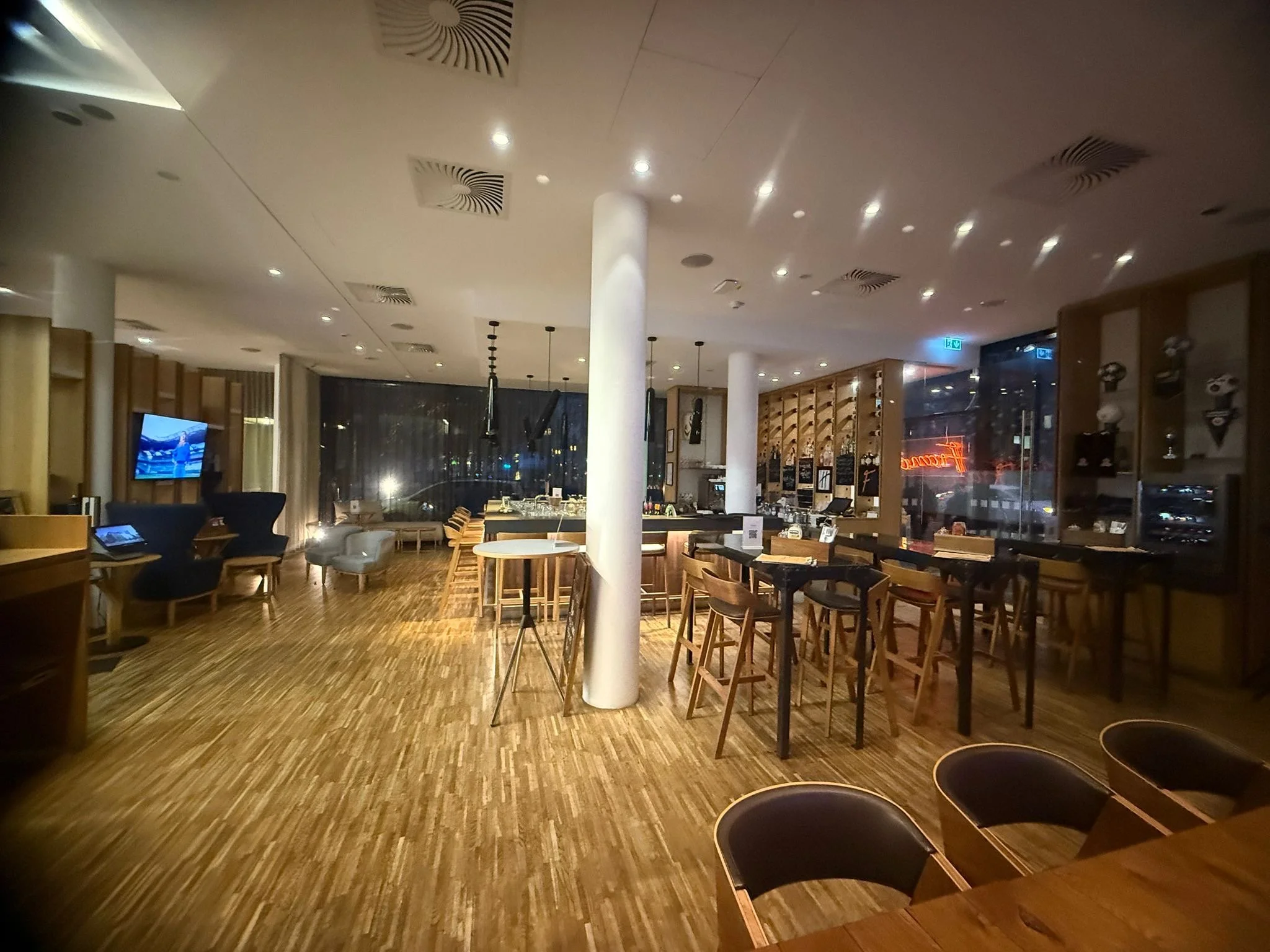 Interior of a modern restaurant or cafe with wooden flooring, a bar area with bar stools, and various seating options including armchairs and high tables. Dim lighting creates a cozy ambiance.