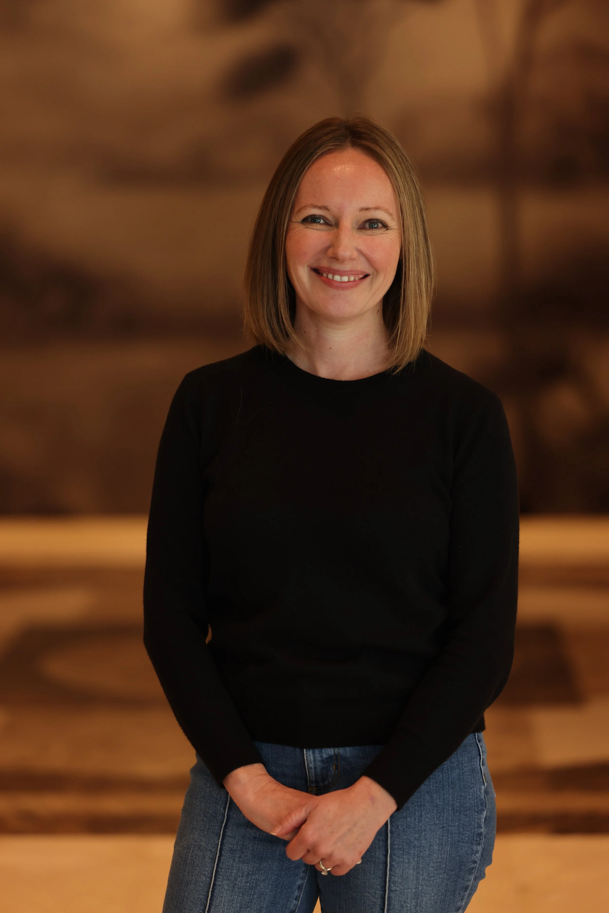 Portrait of a woman with shoulder-length brown hair wearing a black top and blue jeans, standing in front of a blurred wooden background, smiling.