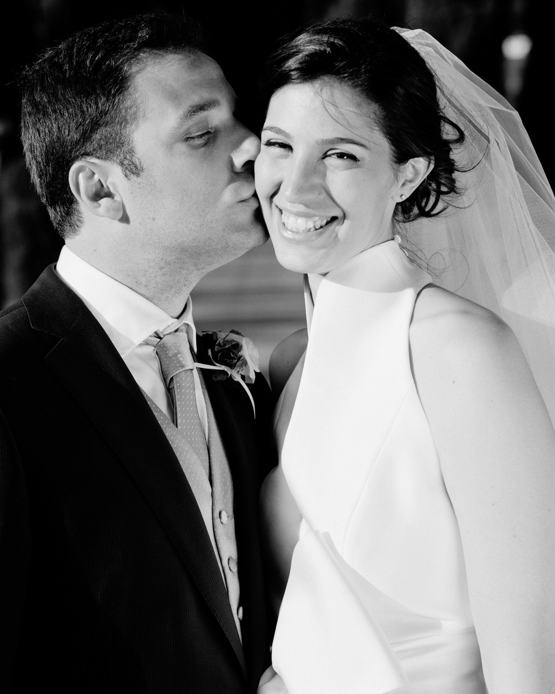 Un uomo e una donna in abiti da matrimonio, lui le sta dando un bacio sulla guancia, mentre lei sorride.