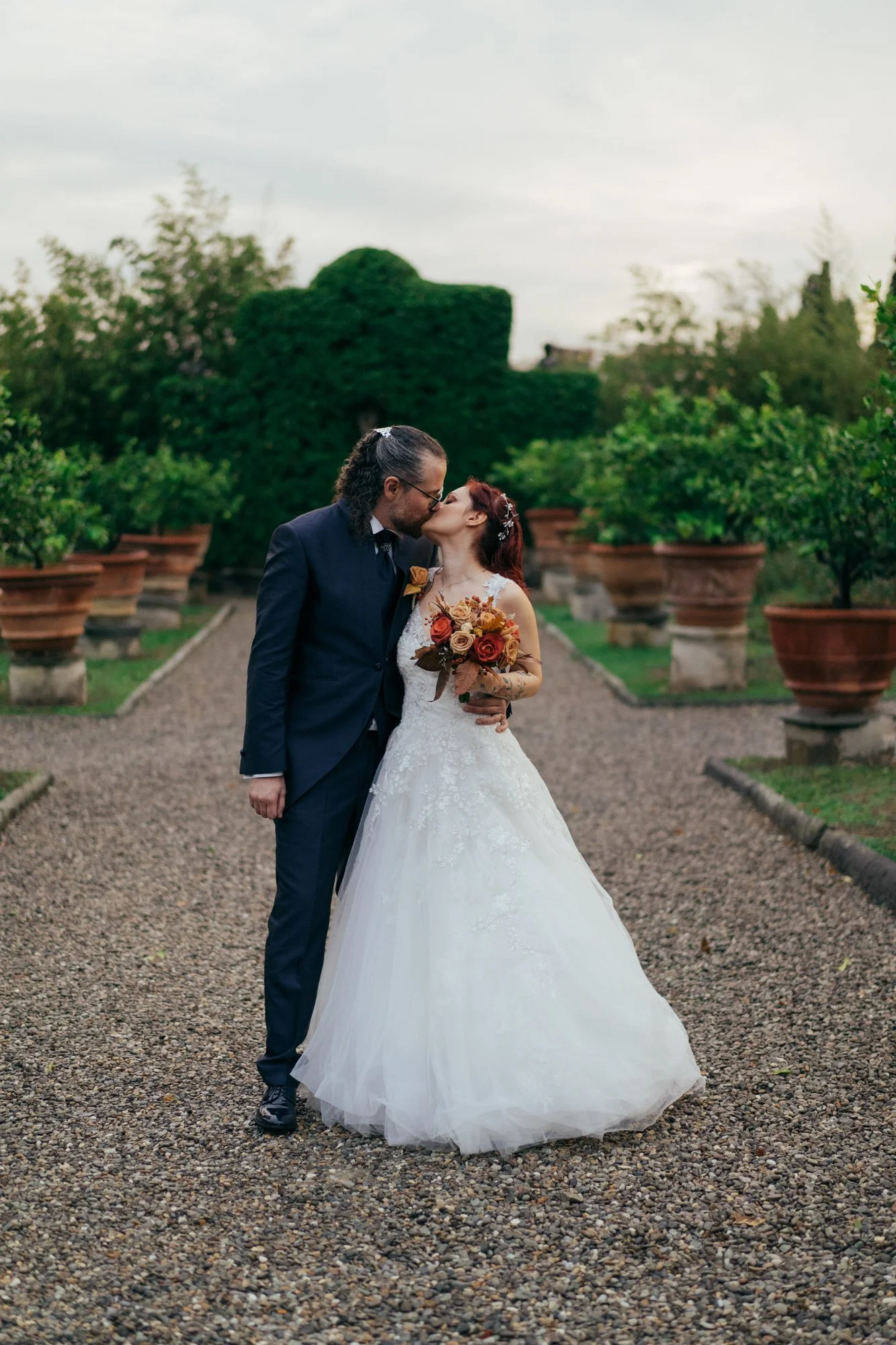 Una coppia di sposi si bacia in un giardino con piante in vaso e cespugli verdi, sotto un cielo nuvoloso.