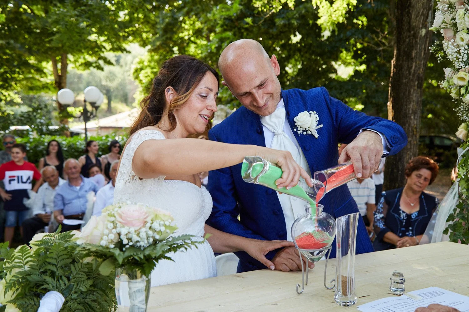 Una coppia di sposi versa sabbia di diversi colori in un angolo di un matrimonio all'aperto, con amici e parenti sullo sfondo.