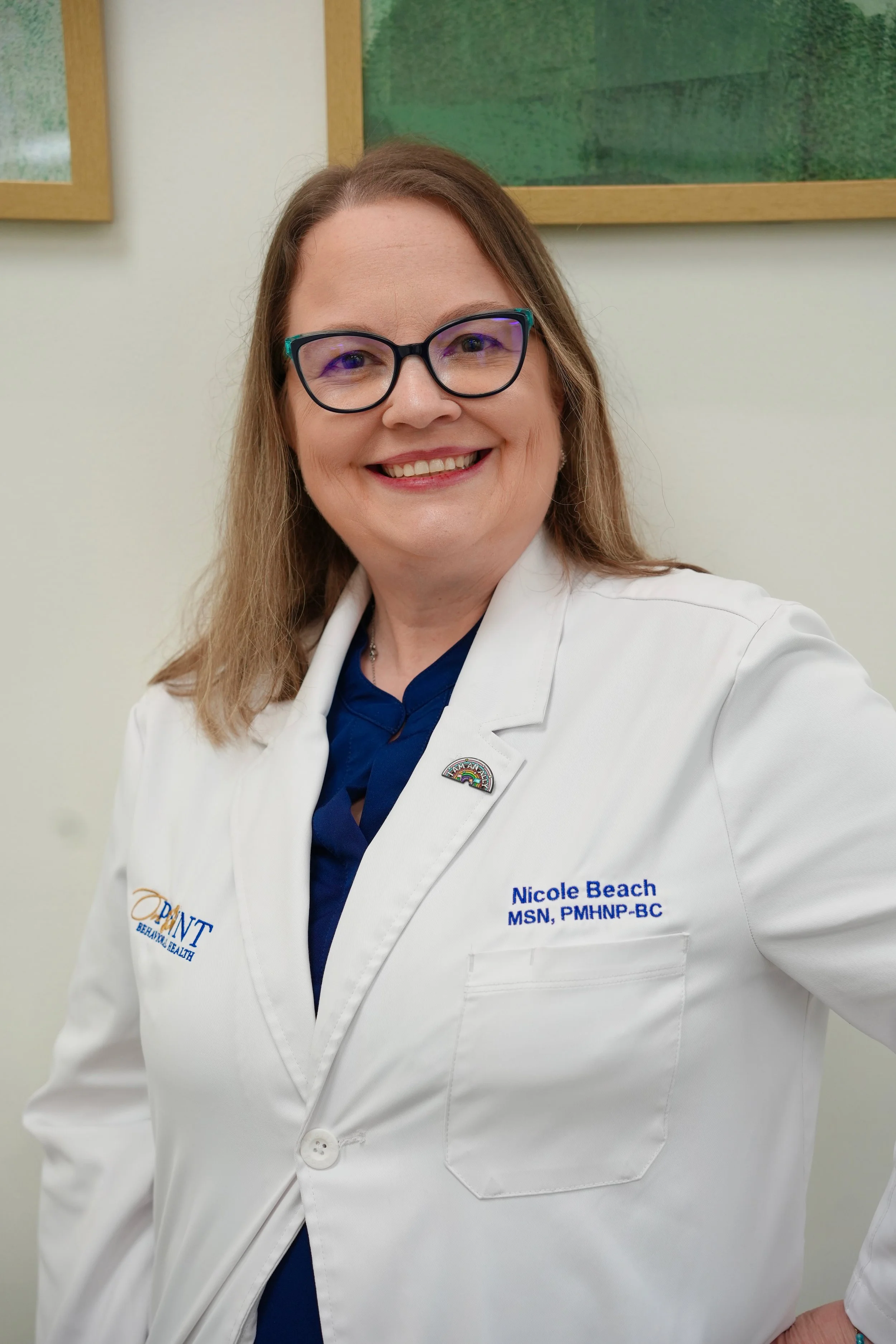 A woman with shoulder-length light brown hair, glasses, and a white medical coat smiling at the camera. She is standing indoors against a beige wall with framed pictures.
