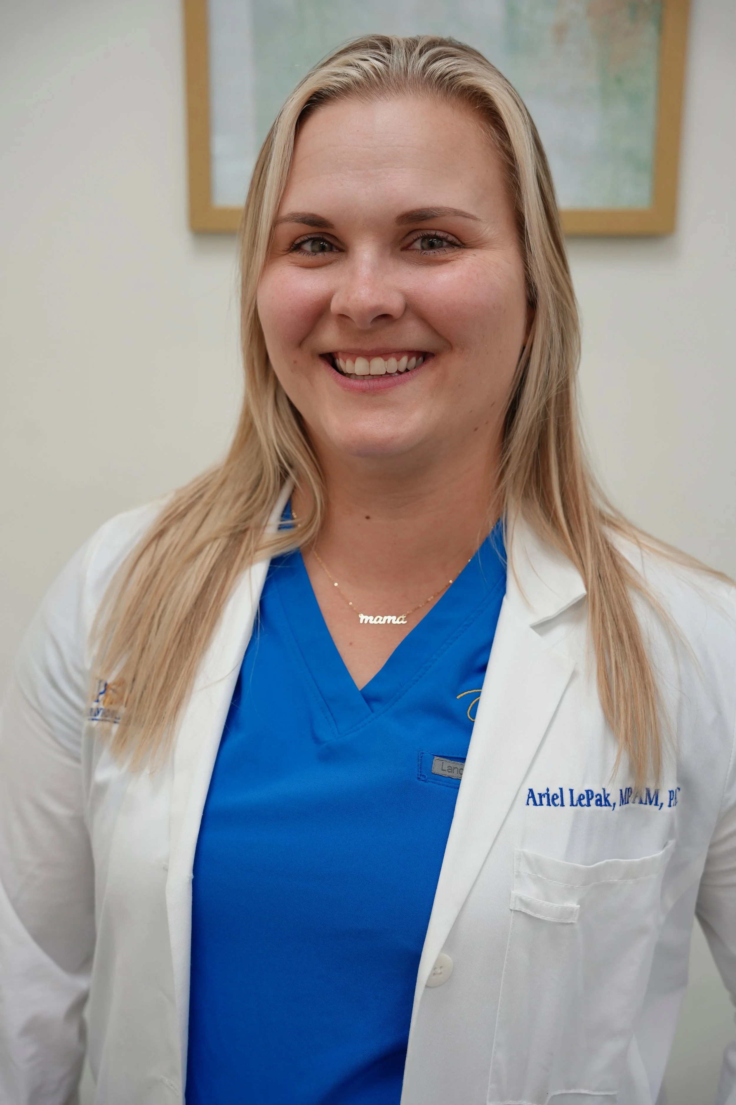 A smiling woman wearing a blue medical uniform with a white lab coat with her name and degree embroidered on it, and a necklace that says 'mama'.