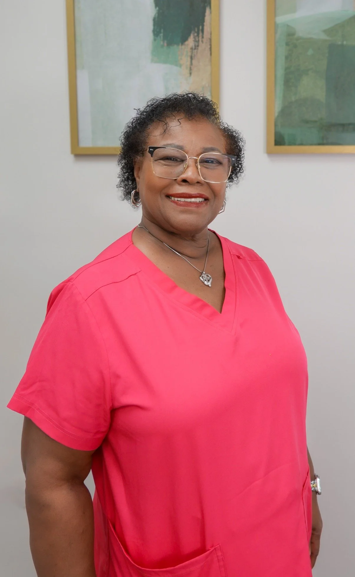 A smiling woman in teal scrubs with glasses and a necklace standing against a white wall with abstract artwork.