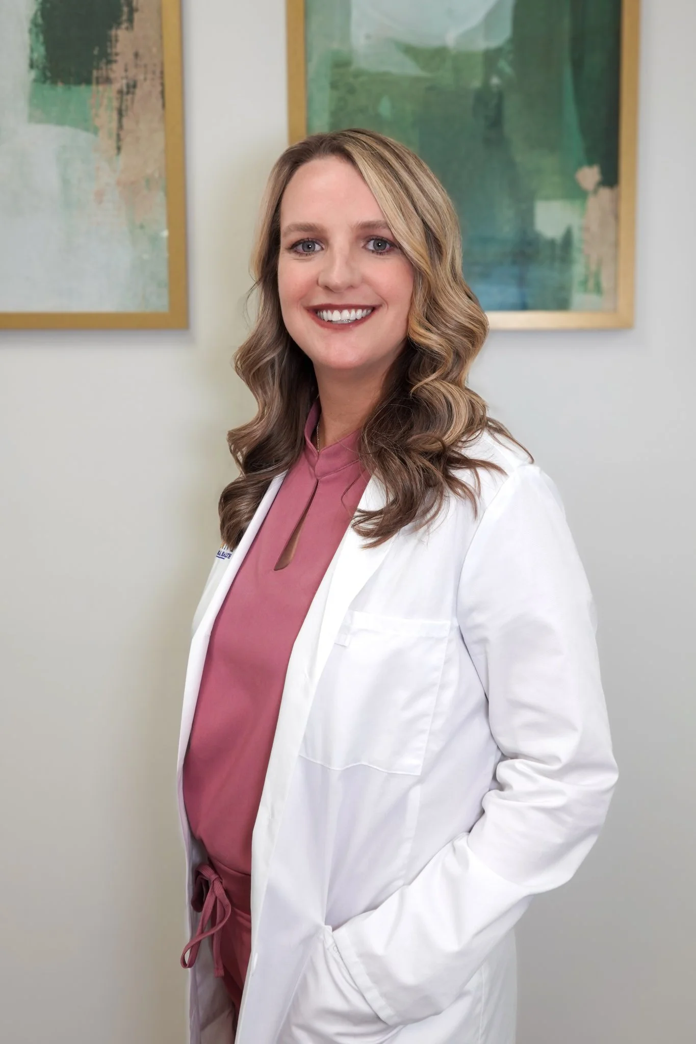 A woman with wavy, shoulder-length hair smiling, wearing a white coat with a logo that reads 'Point Behavioral Health,' standing in front of abstract artwork on a white wall.
