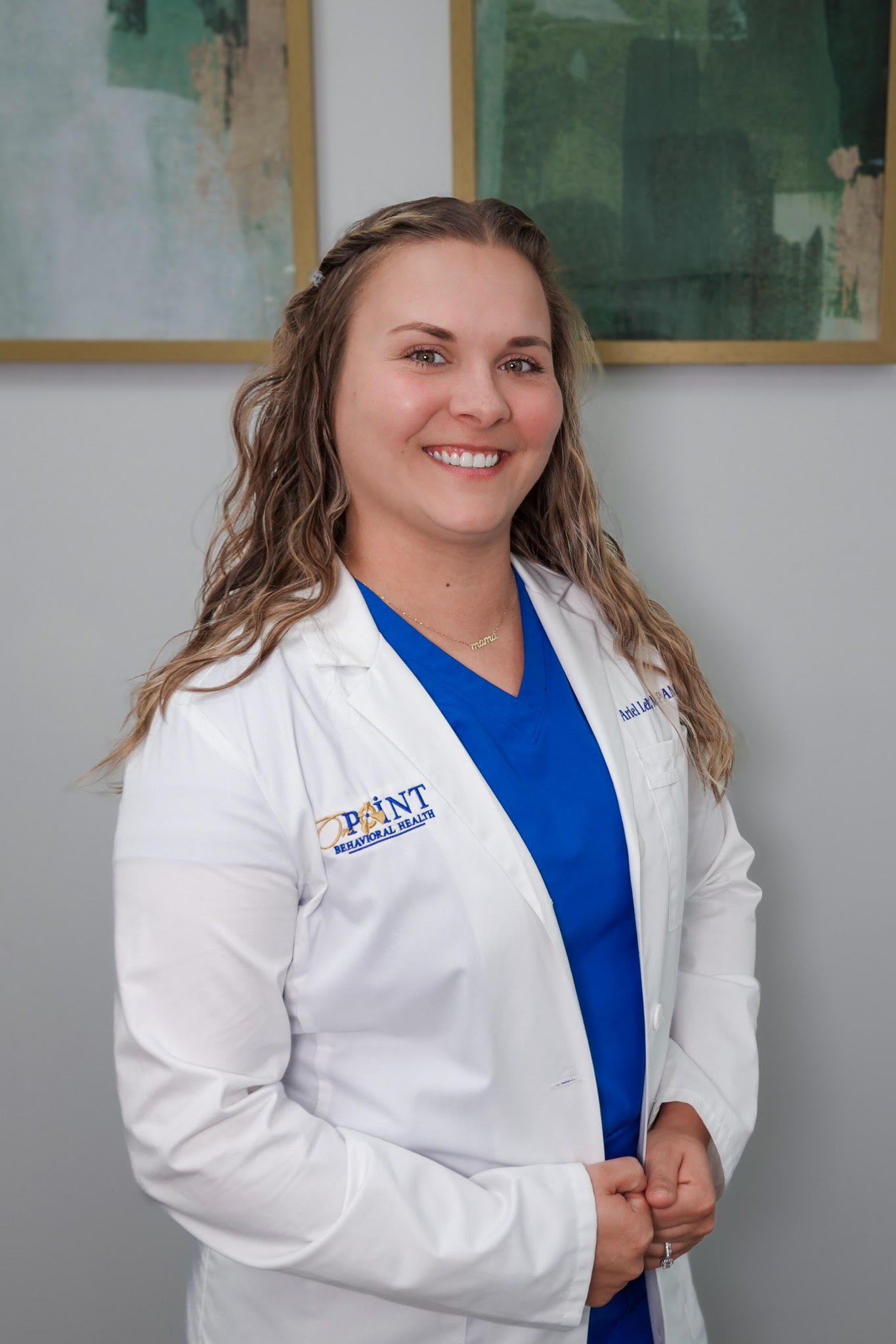 A smiling woman wearing a blue medical uniform with a white lab coat with her name and degree embroidered on it, and a necklace that says 'mama'.