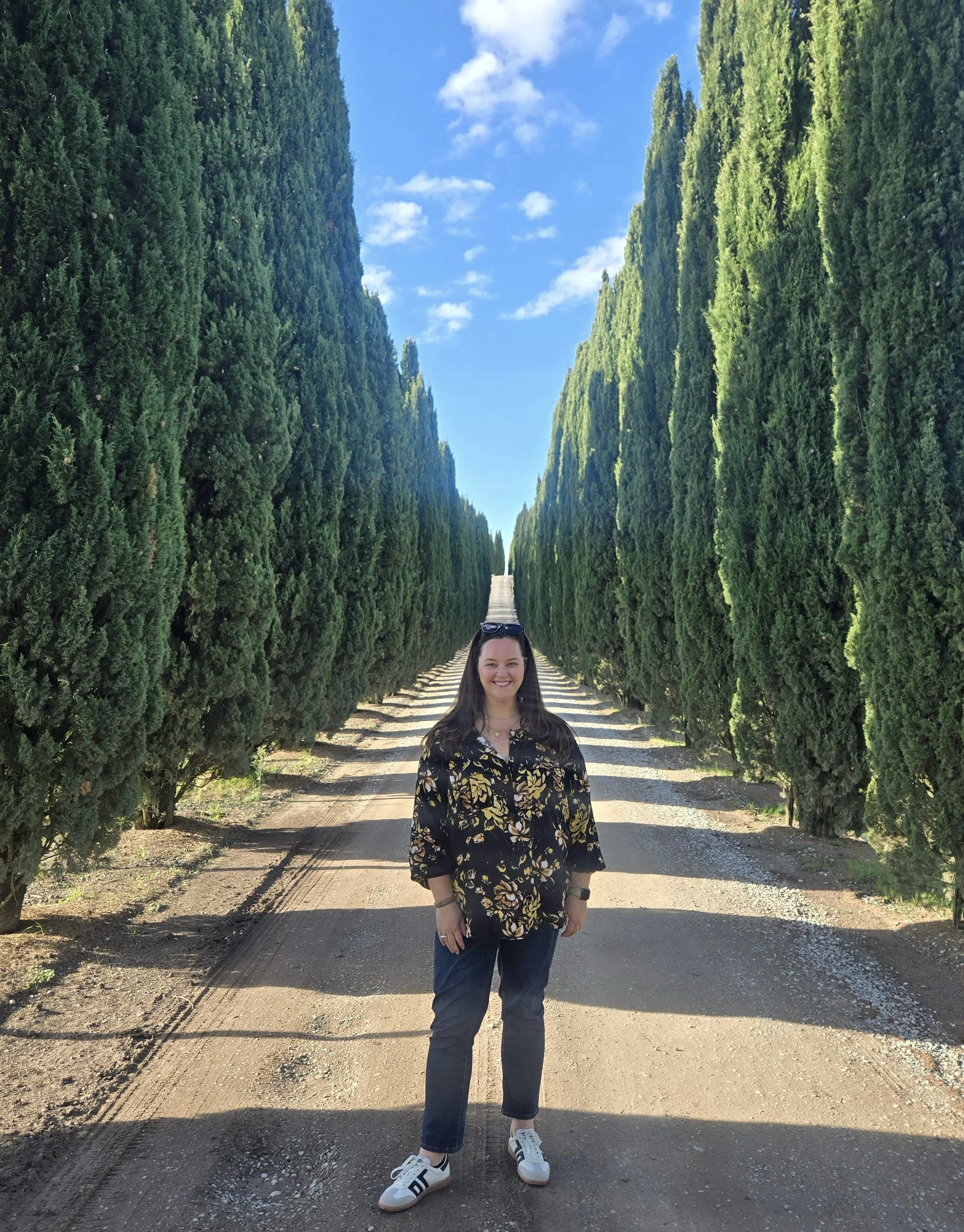 A woman standing on a dirt road lined with tall, lush green cypress trees on both sides, with a blue sky and scattered clouds overhead.
