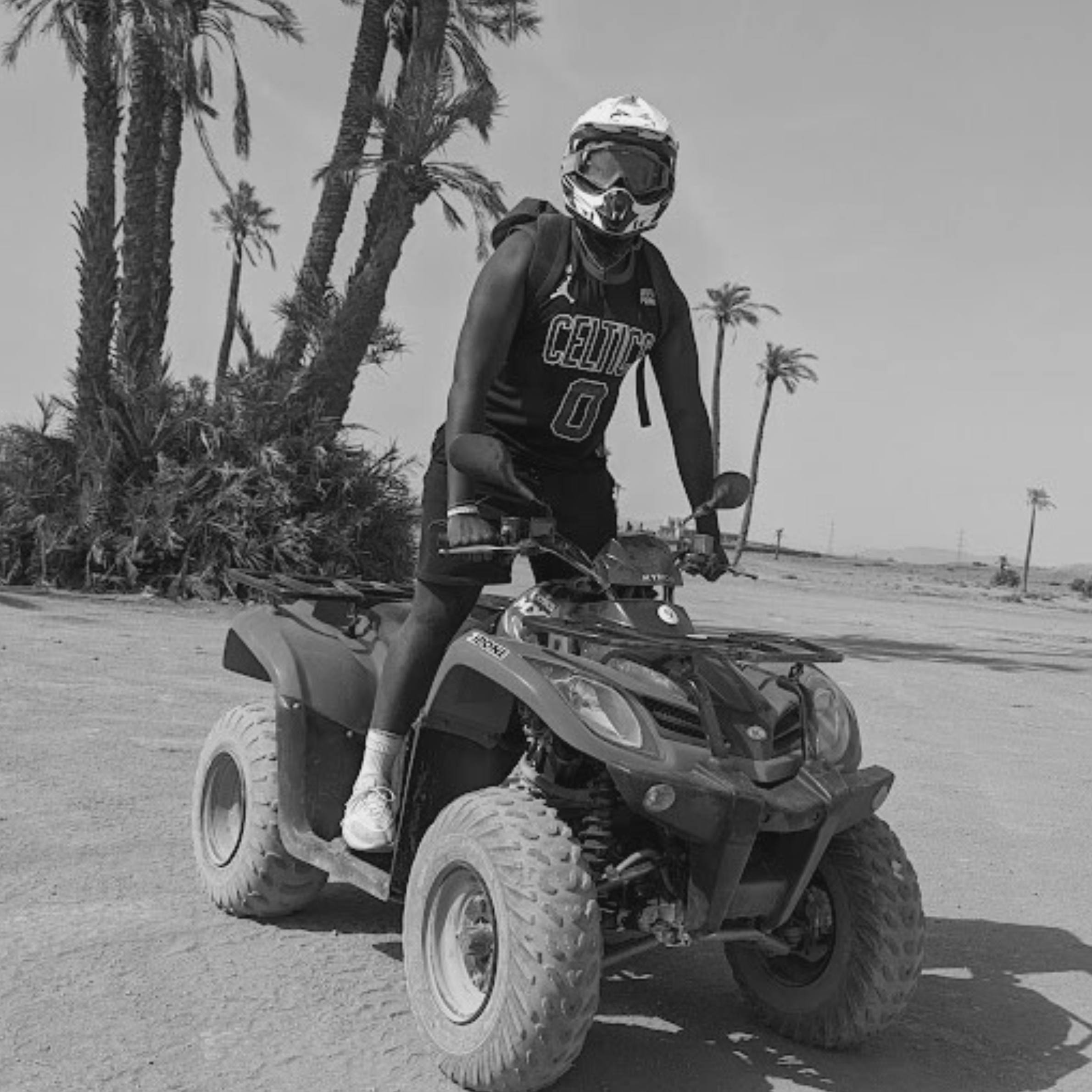 Mark wearing a helmet, sunglasses, and a Boston Celtics jersey riding an all-terrain vehicle in a desert area with palm trees.
