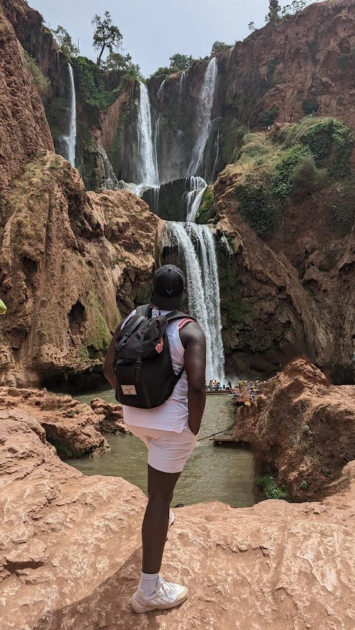 Mark wearing a backpack and a cap standing on rocky terrain, facing a large waterfall with multiple streams falling into a pool below.