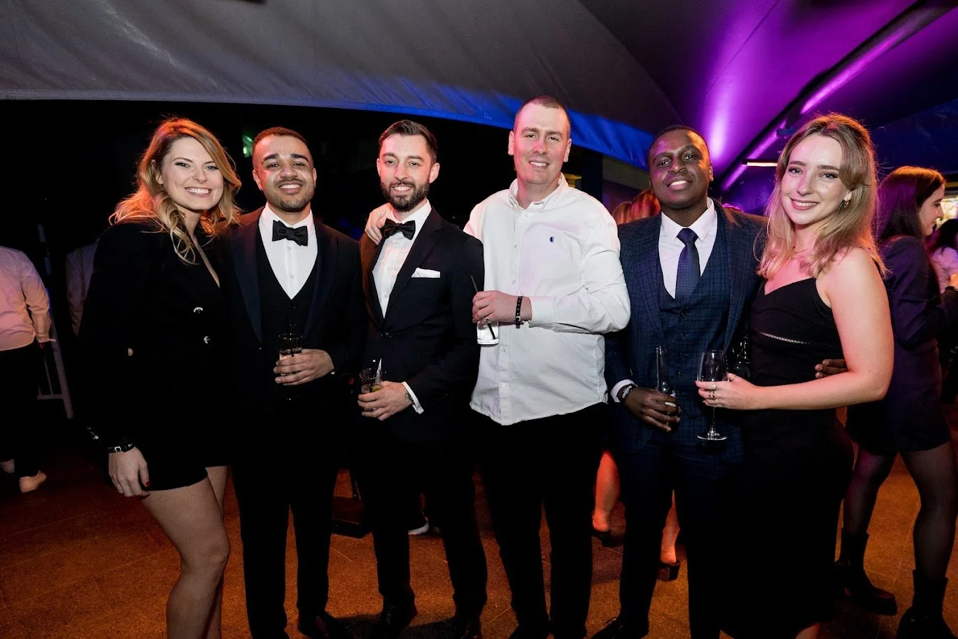 Group of six people dressed in formal attire at a party or event, posing together and smiling.