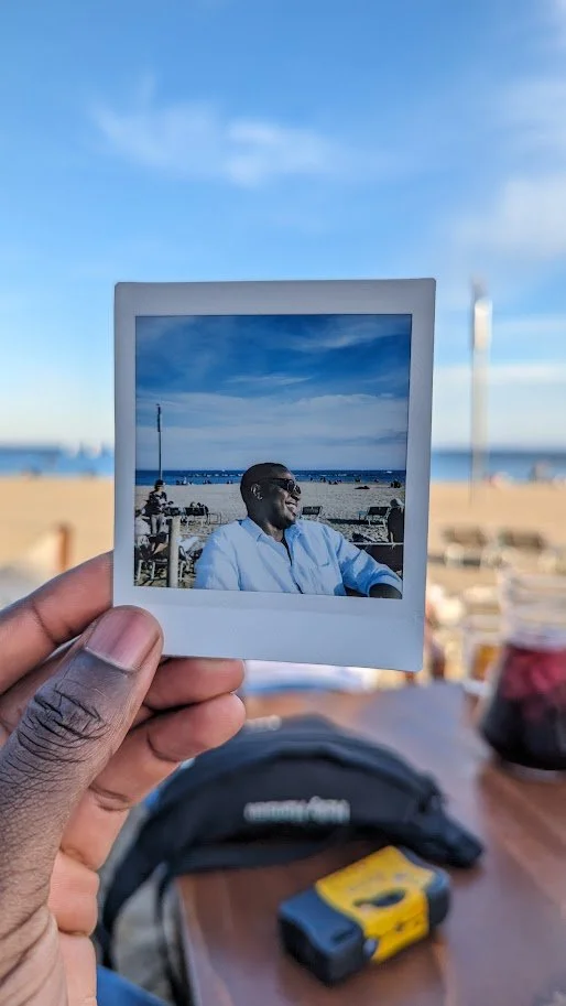A person holding a Polaroid photo of a smiling man in a white shirt sitting outdoors at the beach, with a blue sky and ocean in the background.