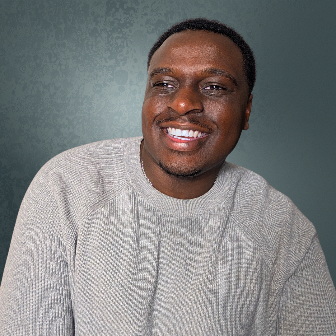 Smiling man with short black hair wearing a light gray sweater, against a textured gray background.