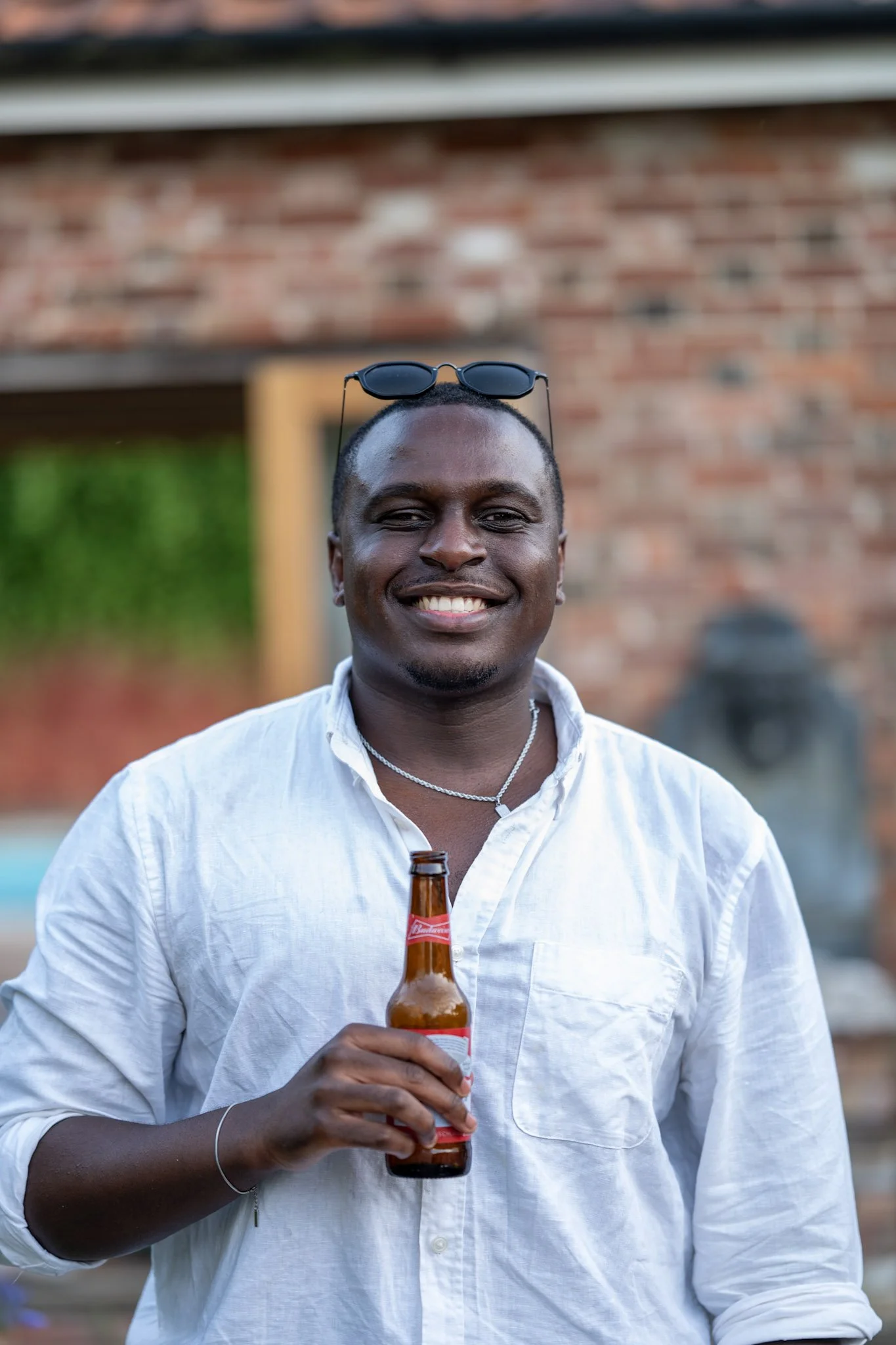A smiling man wearing a white shirt, holding a beer bottle, with sunglasses on his head, standing outdoors with a brick wall and greenery in the background.