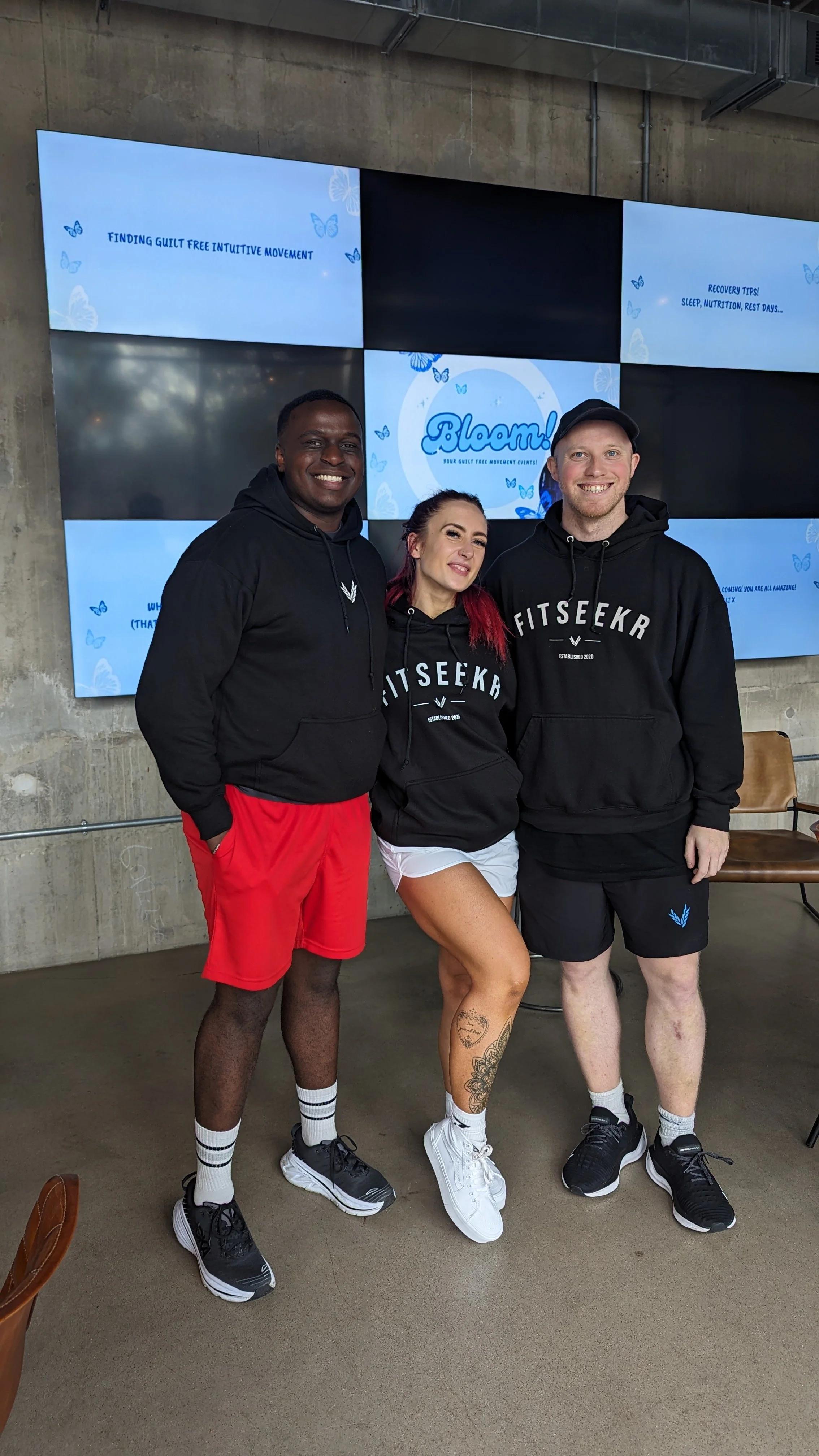 Three people in athletic attire standing together indoors in front of television screens showing event graphics. They are smiling and wearing matching hoodies with "FITSEEKR" printed on them.