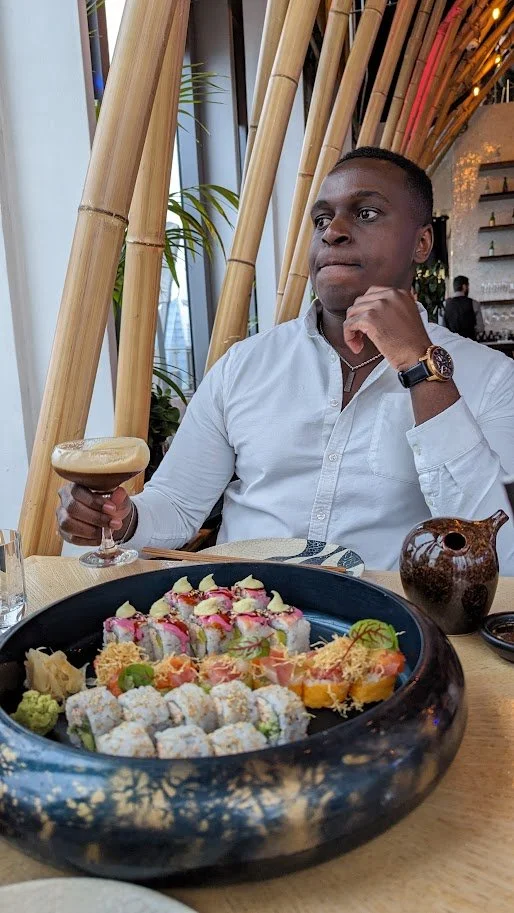 A man sitting at a restaurant table with a large platter of sushi, holding a cocktail in a coupe glass, in a modern restaurant with bamboo decor.