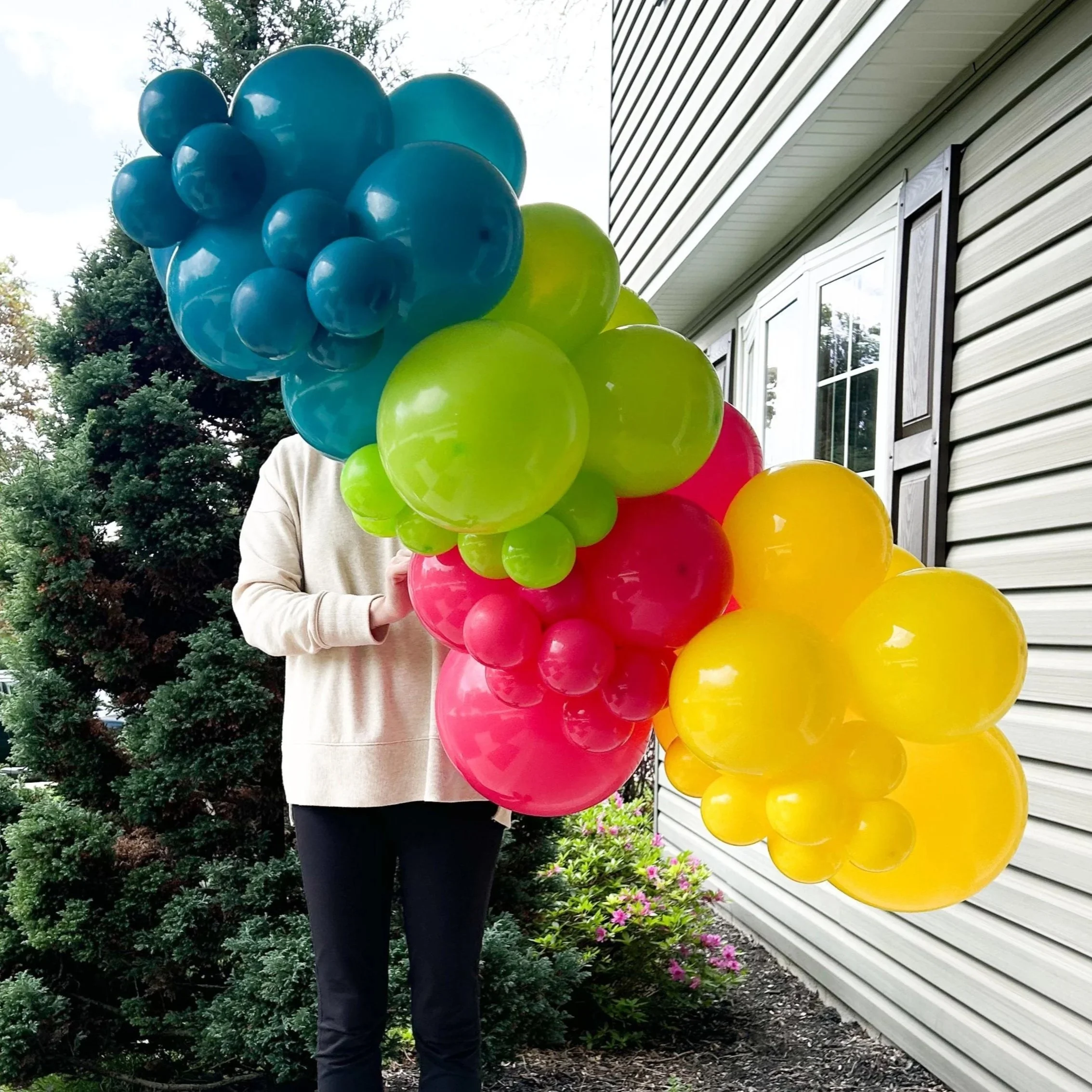 Person holding colorful balloons in front of a house and bushes.