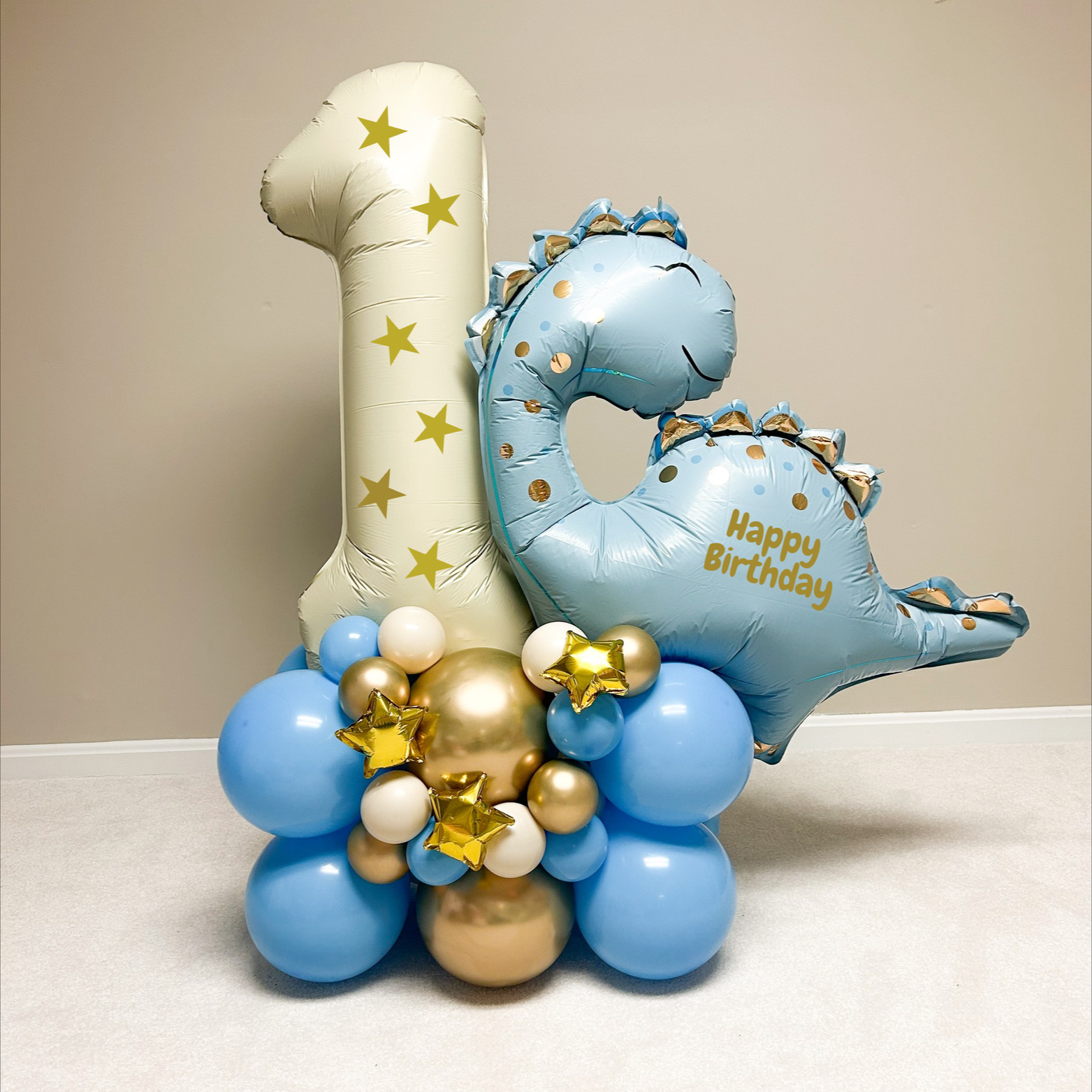 Balloon arrangement with a large number 1, a dinosaur-shaped balloon with "Happy Birthday" written, and various blue, white, and gold balloons.