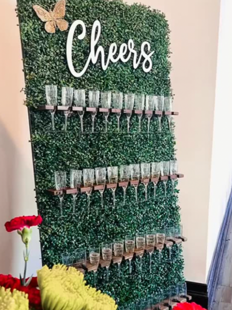 A decorative wall with the word 'Cheers' at the top, adorned with artificial greenery, and holding multiple rows of champagne glasses filled with drinks.