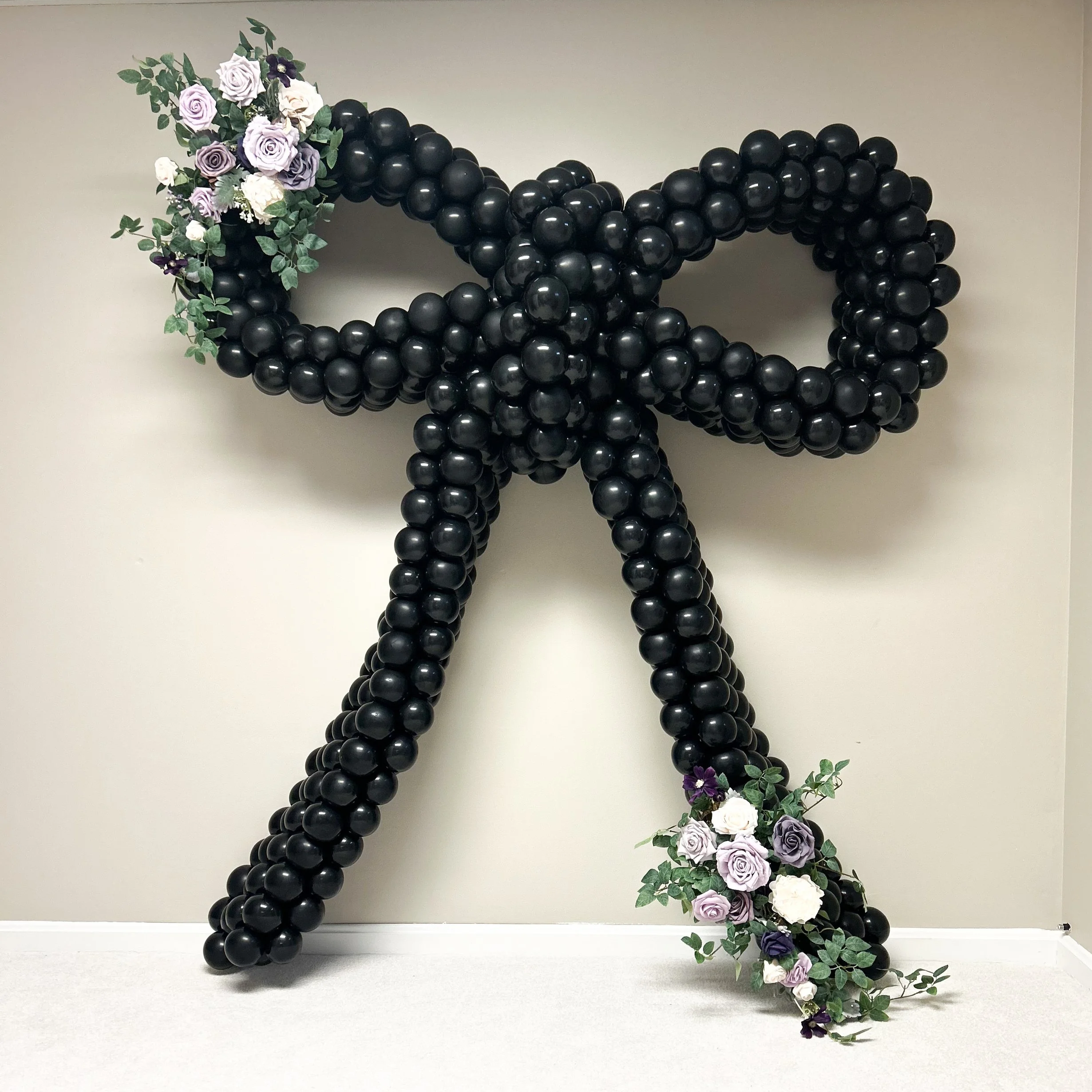 Black balloon sculpture shaped like a bow with flower arrangements at the ends, featuring purple, white, and pale violet roses with green leaves.