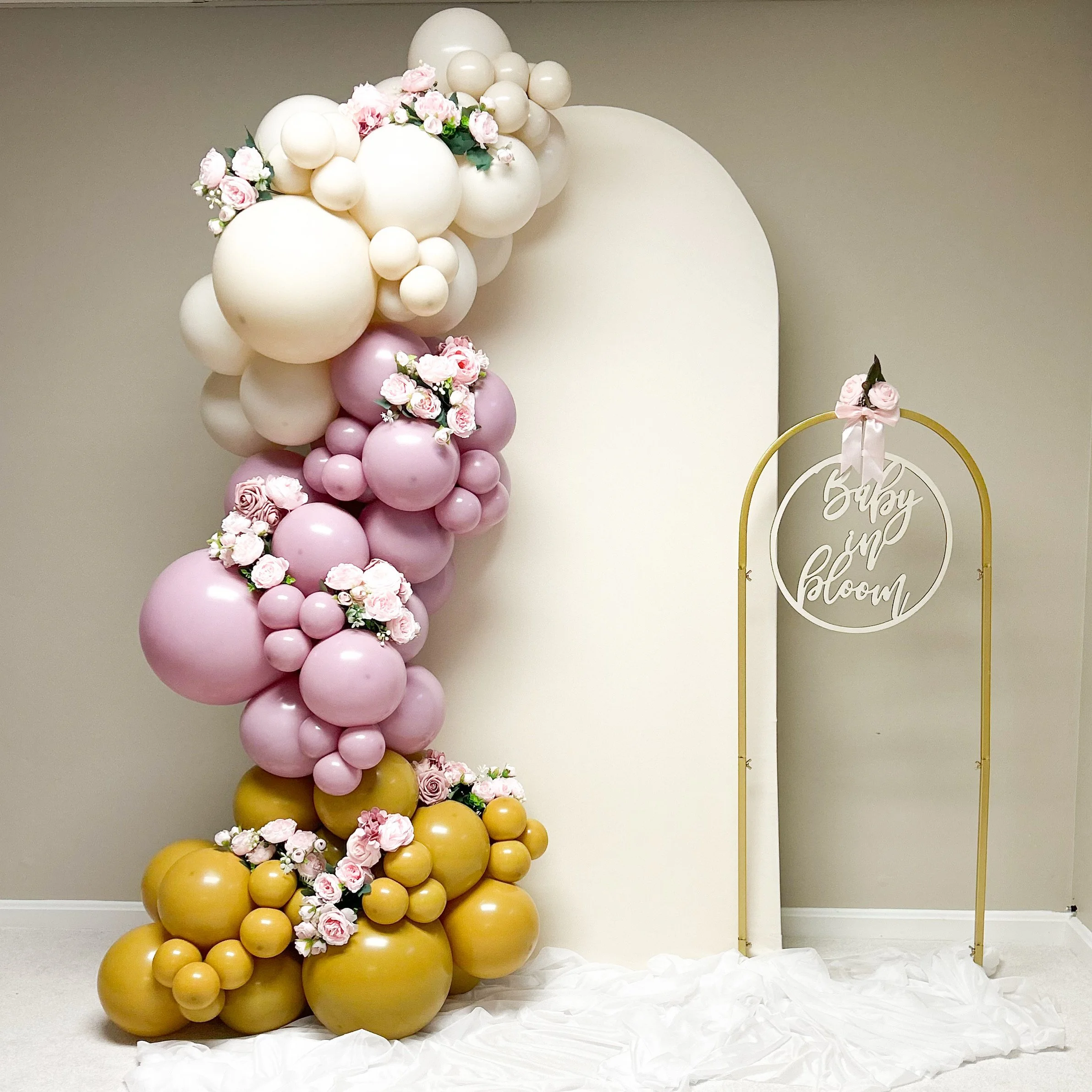 Baby shower balloon arrangement with pink and gold balloons, pink flowers, and a white backdrop with a gold frame that has a circular sign reading 'Stay and Bloom' in cursive writing in Cleveland, Northeast Ohio.