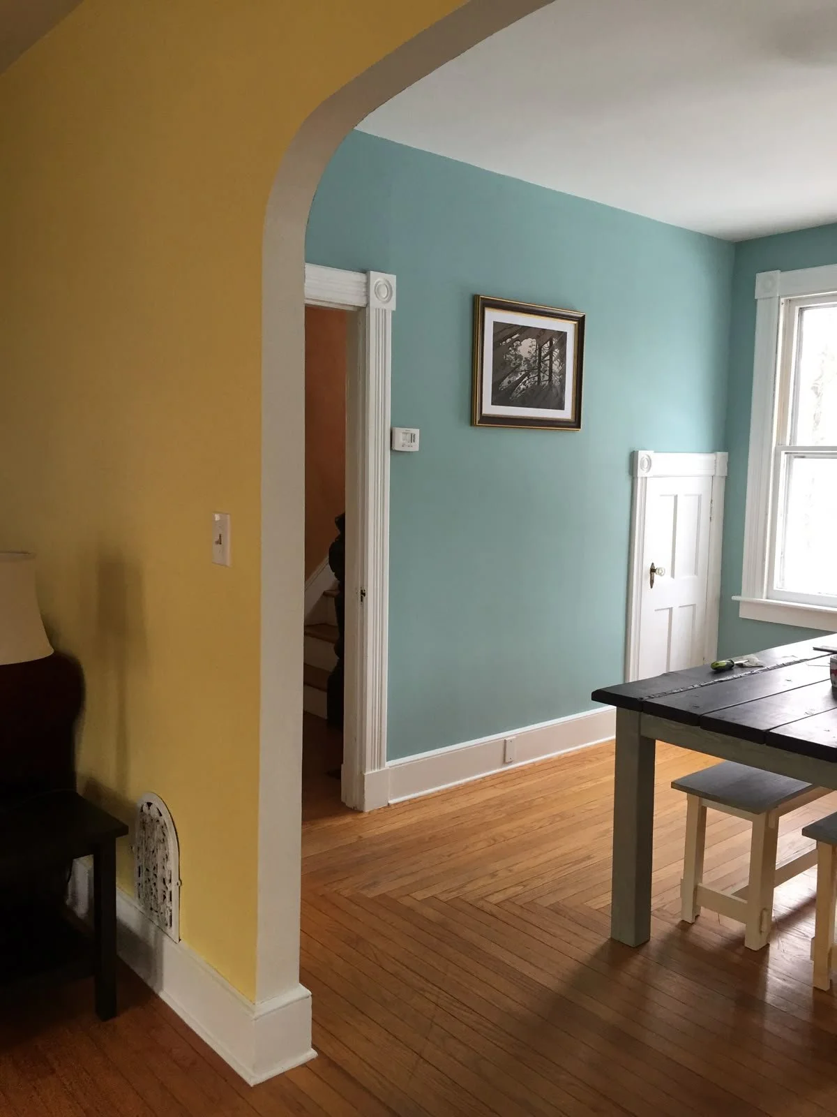 Interior painting, home improvement paint project, cozy spaces. Interior view of a room with wooden flooring, light blue and yellow walls, a window, a framed black-and-white picture, and a small door built into the wall.