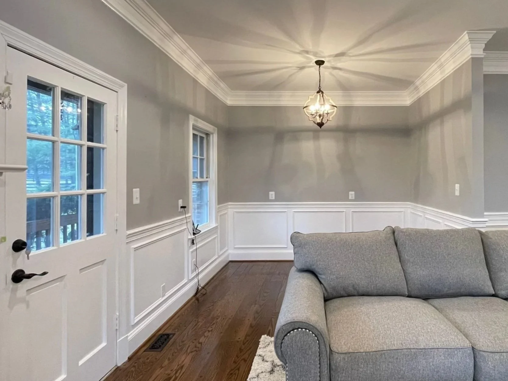 Residential home interior living room freshly painting with gray walls, white wainscoting, a chandelier, two windows, a nearby door with glass panes, hardwood floor, and a gray sectional sofa.