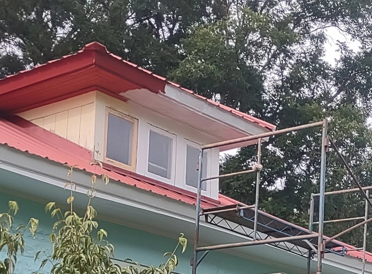 A house with a red metal roof, partially painted walls, and a scaffolding structure outside.