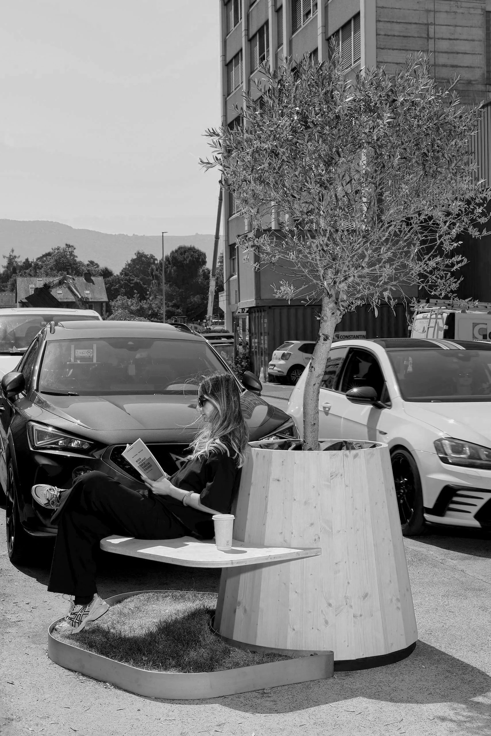 A woman reading on a custom modular wooden bench in a public urban space, illustrating the integration of contemporary street furniture into the city environment.