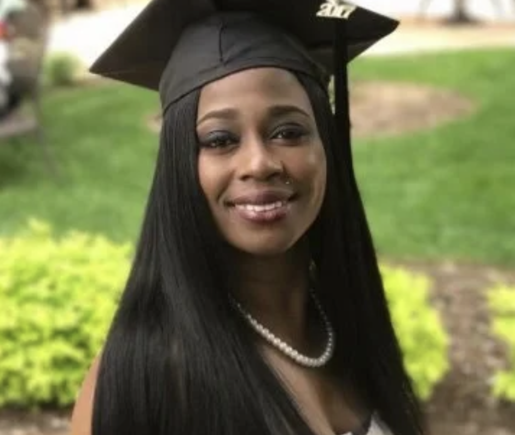 Woman wearing graduation cap and gown, smiling, outdoors in a park.