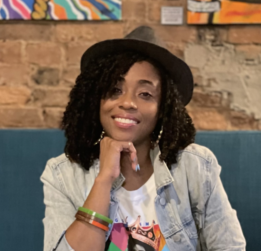 A woman sitting on a teal bench with a painted brick wall in the background. She has curly hair, wears a black hat, a light denim jacket, and colorful bracelets. She is smiling and resting her chin on her hand.