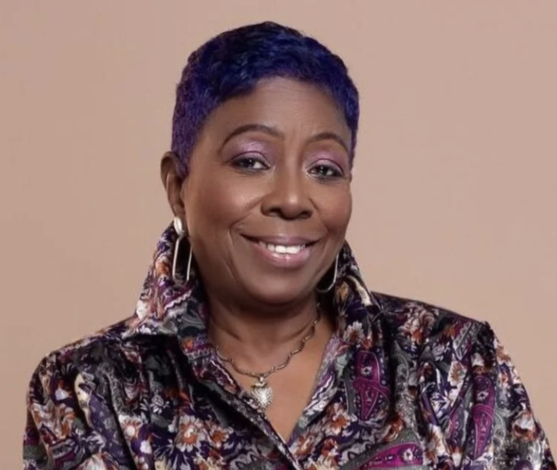 A woman smiling with short dark purple hair, wearing a patterned blouse, hoop earrings, and a necklace.