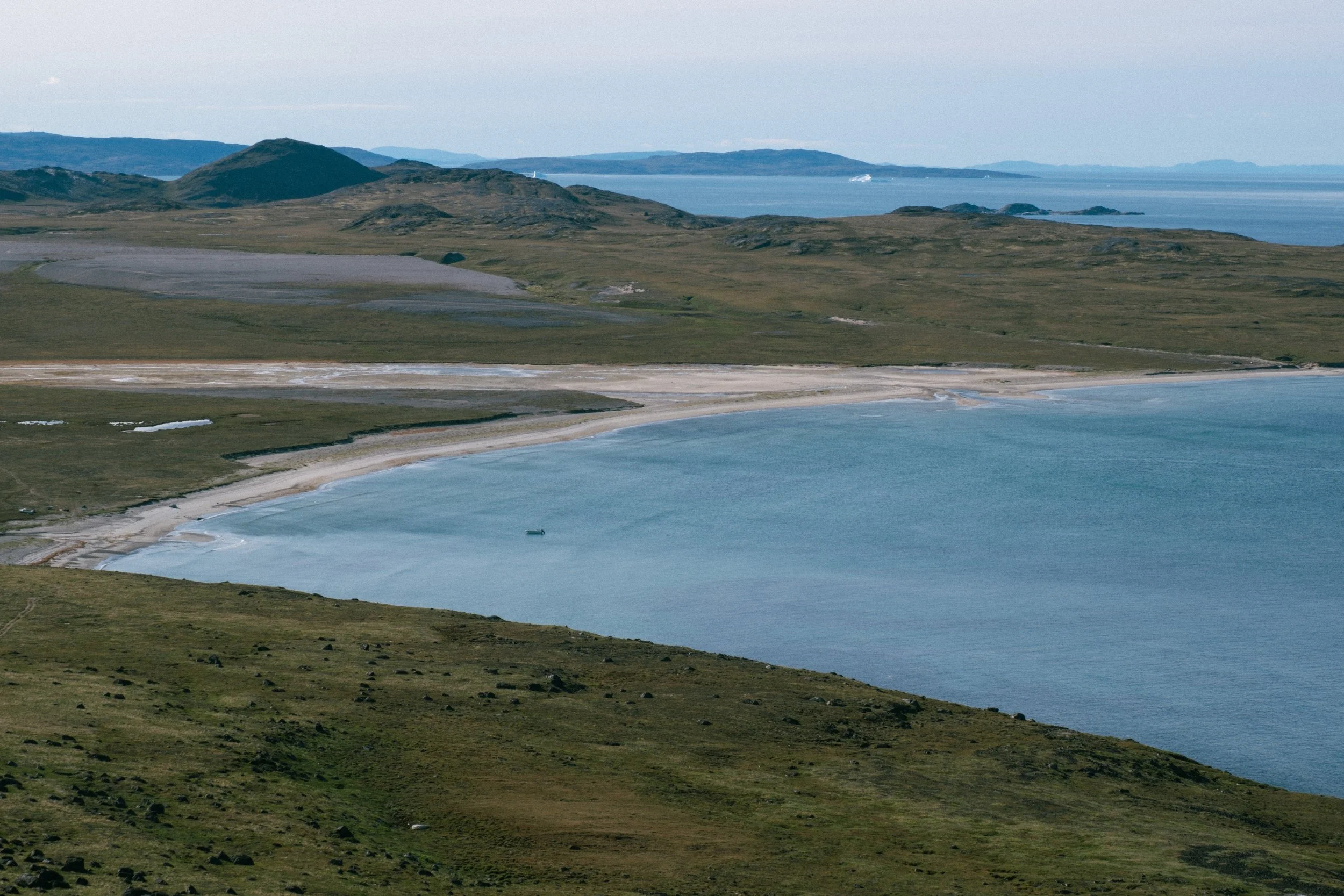 Scenic landscape with green hills, a sandy shoreline, and a large body of water, possibly a bay or ocean, in the background, under a clear sky.