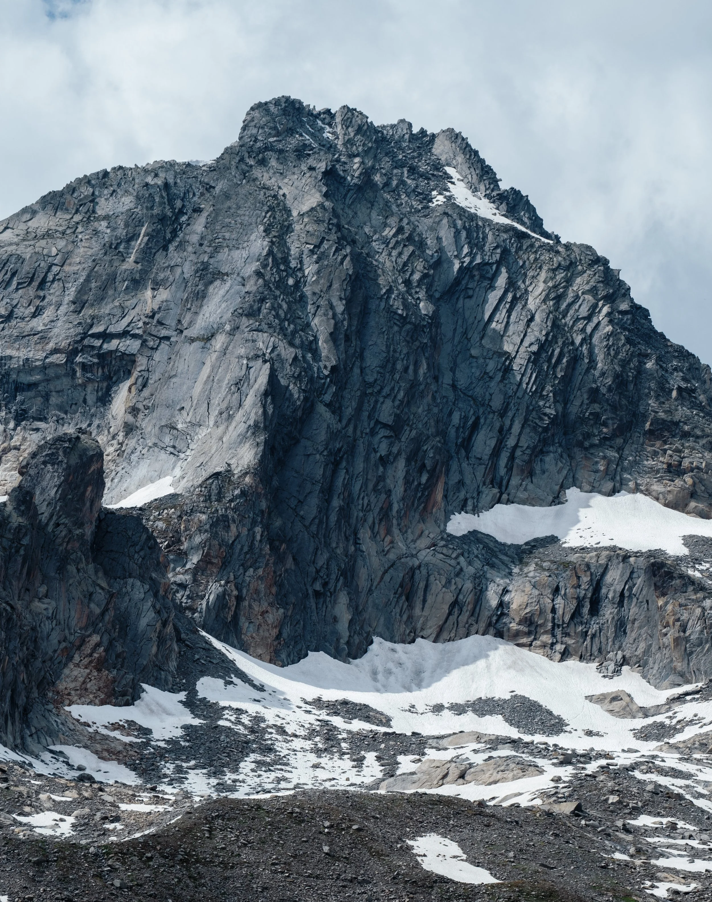 A rugged mountain landscape with sharp, rocky peaks and patches of snow at the base and on the slopes, under a cloudy sky.