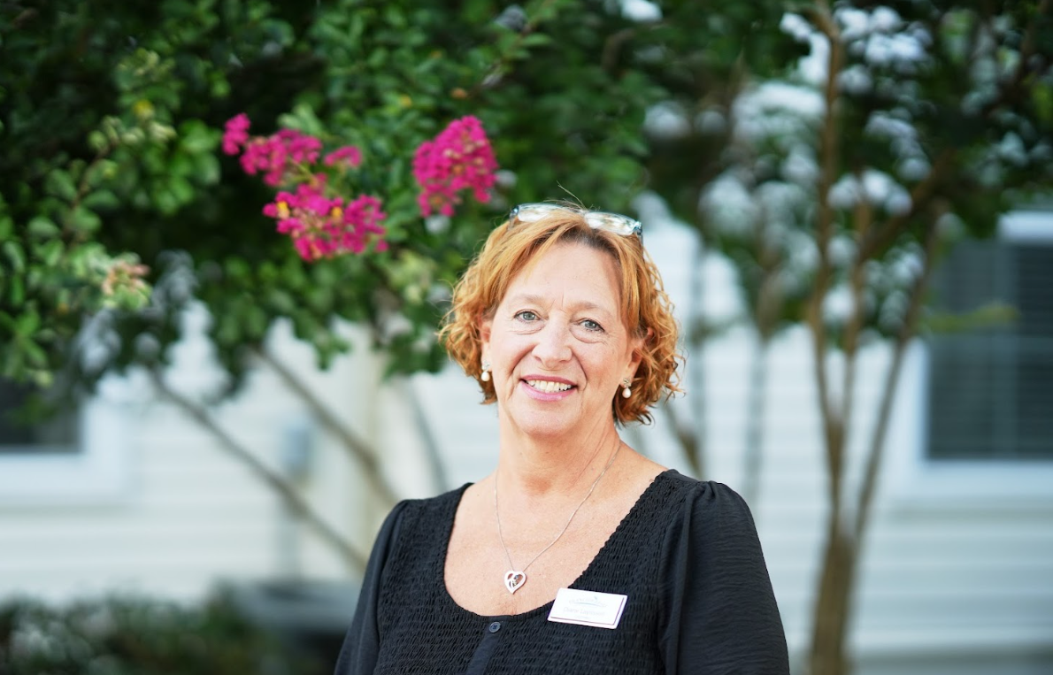 A smiling woman with red curly hair, wearing a black top, glasses on her head, earrings, and a name badge.