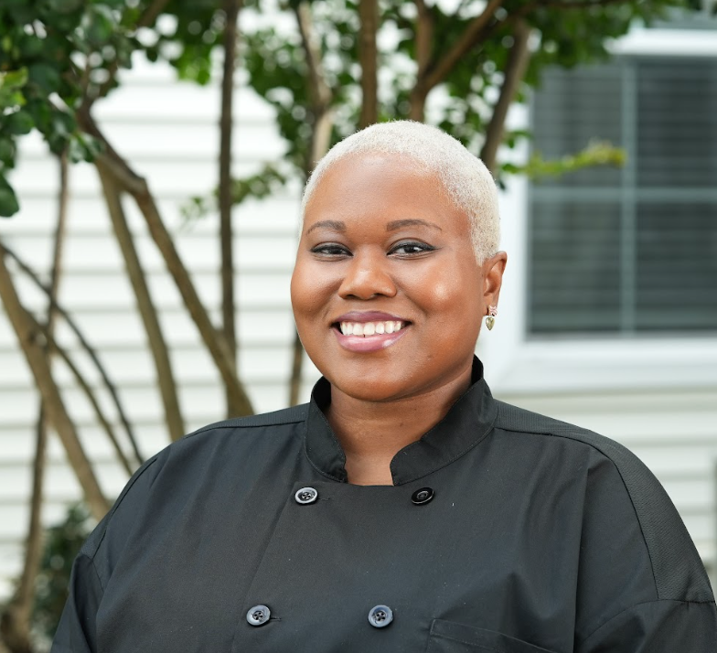 A smiling woman with short hair, wearing glasses, gold hoop earrings, and a black top, standing against a plain light-colored background.