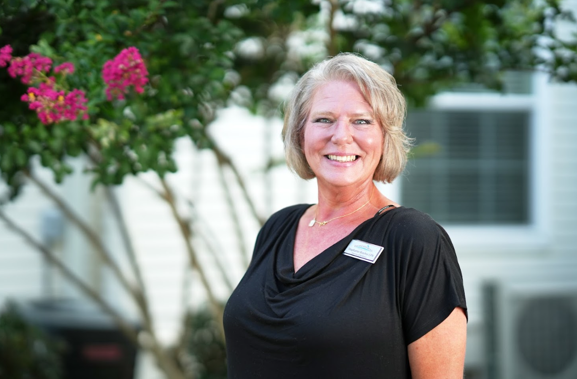 A woman with shoulder-length gray hair and glasses, smiling, wearing a black jacket with the logo 'François Bend Senior Living' on it, standing against a plain off-white wall.