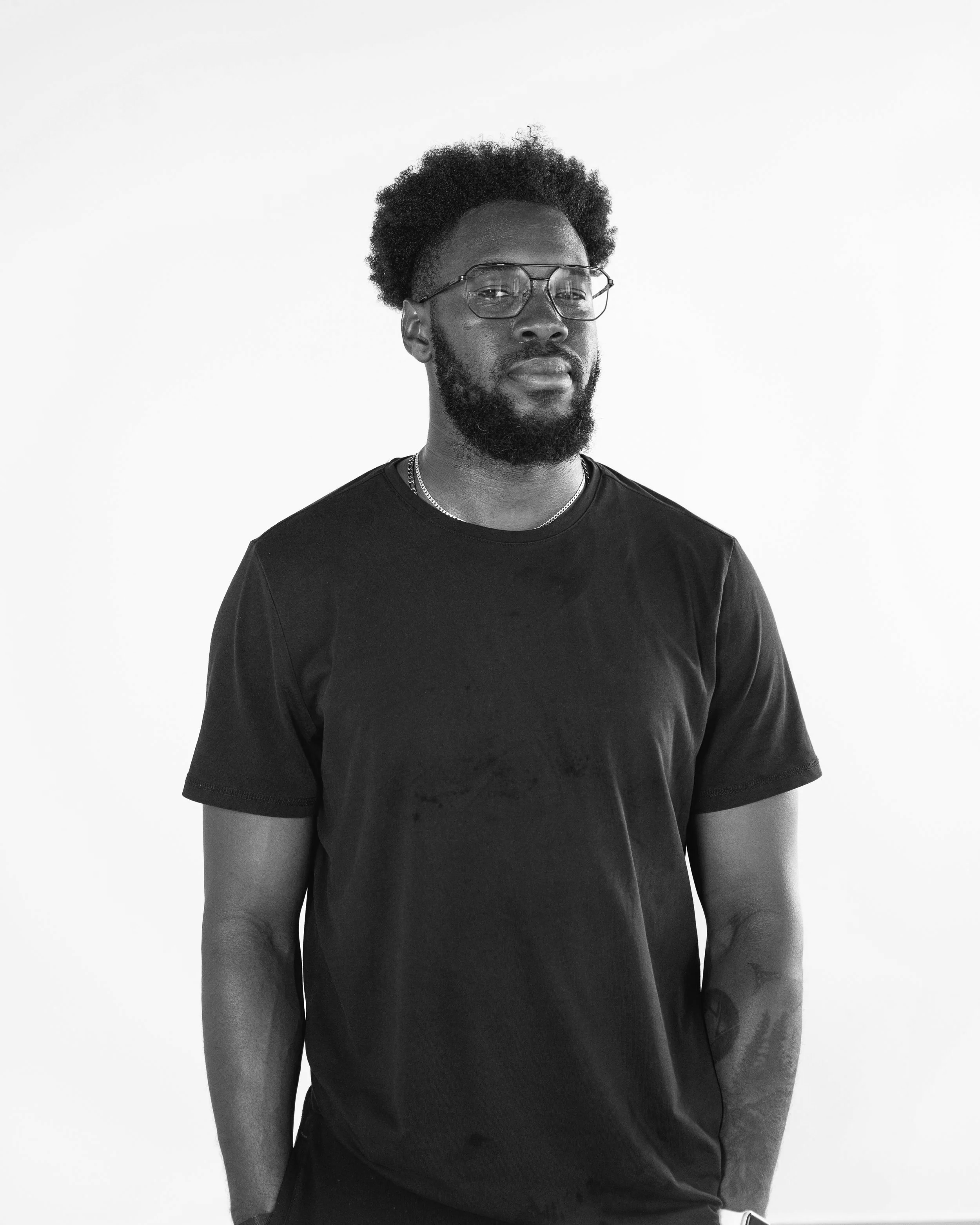 Black and white portrait of a man with glasses, a beard, and curly hair, wearing a plain dark T-shirt against a plain light background.