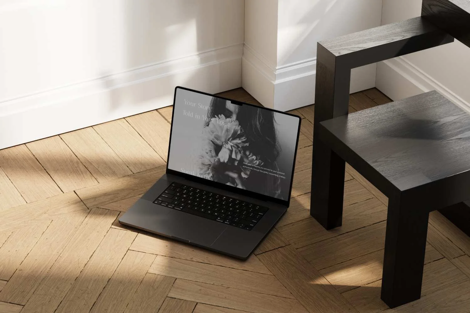 A laptop on the floor next to a black wooden table, with sunlight casting shadows on a herringbone wooden floor and a white wall.