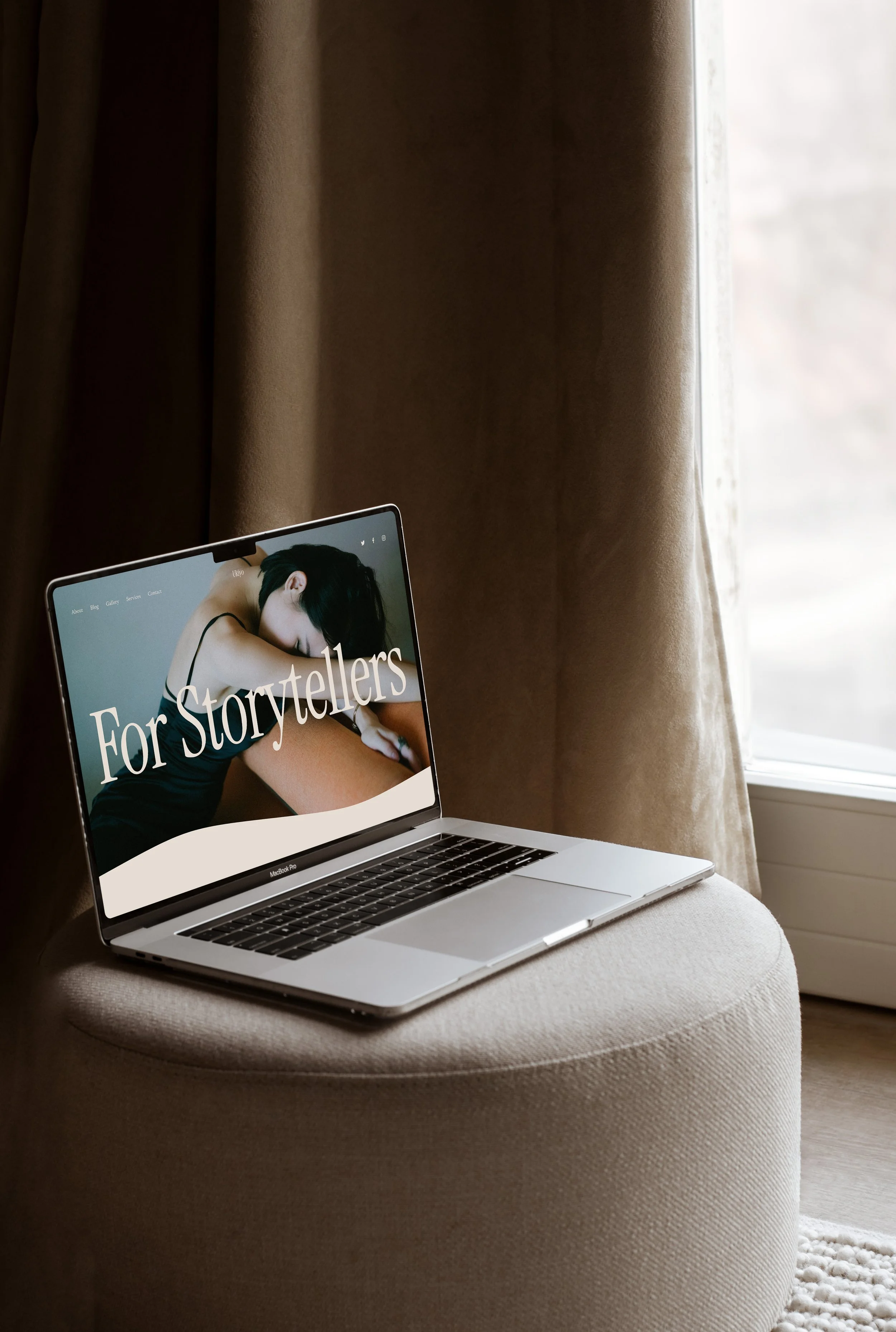 A silver MacBook Pro laptop on a beige cushioned bench, displaying a website with a woman in a black tank top and the words "For Storytellers" on the screen. The scene is next to a window with beige curtains letting in natural light.