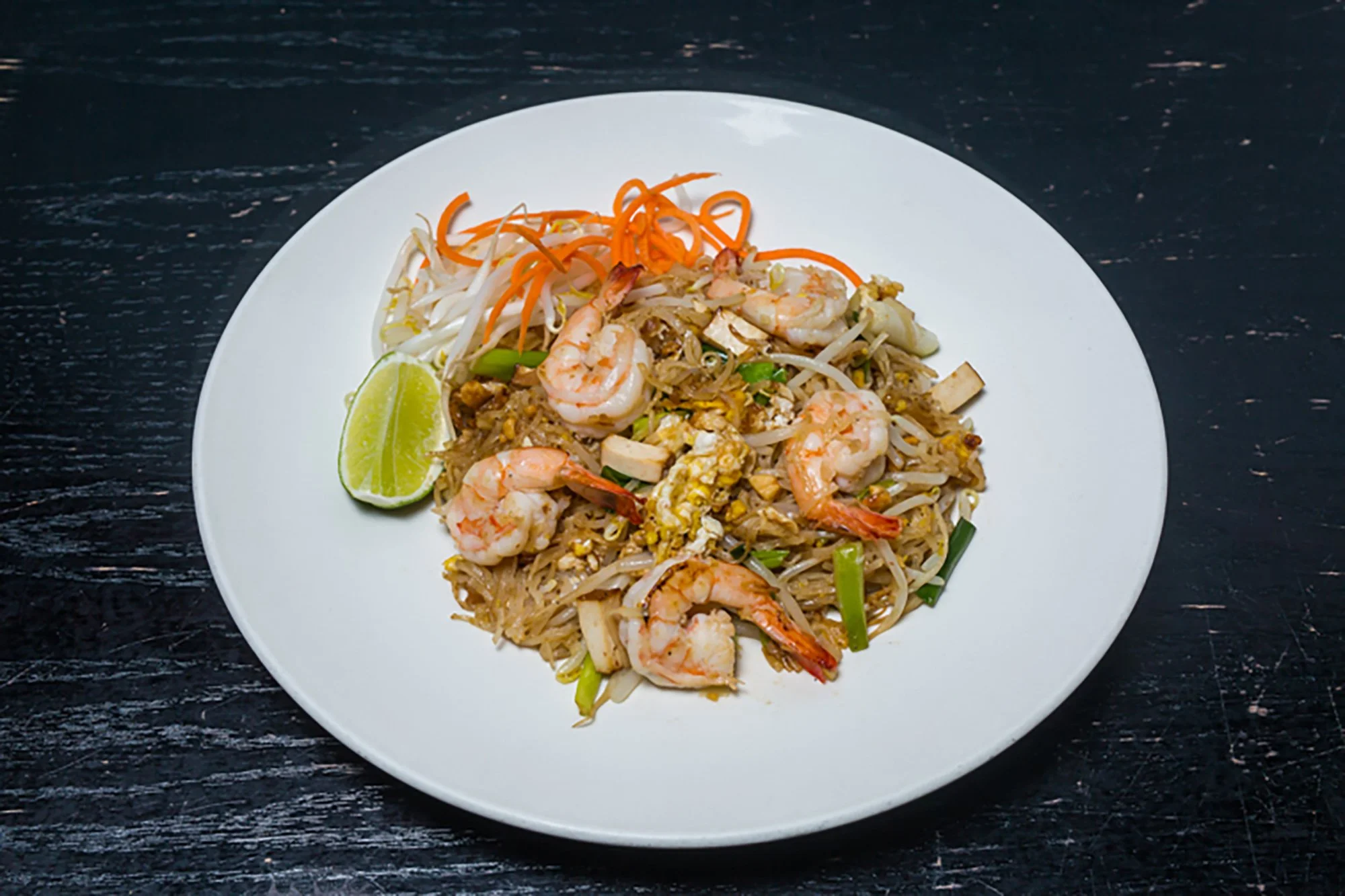 Plate of shrimp pad thai with lime wedge, bean sprouts, and shredded carrots on black wooden table.