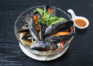 Bowl of cooked mussels with fresh basil leaves and orange chili peppers, served with a small dish of red dipping sauce.
