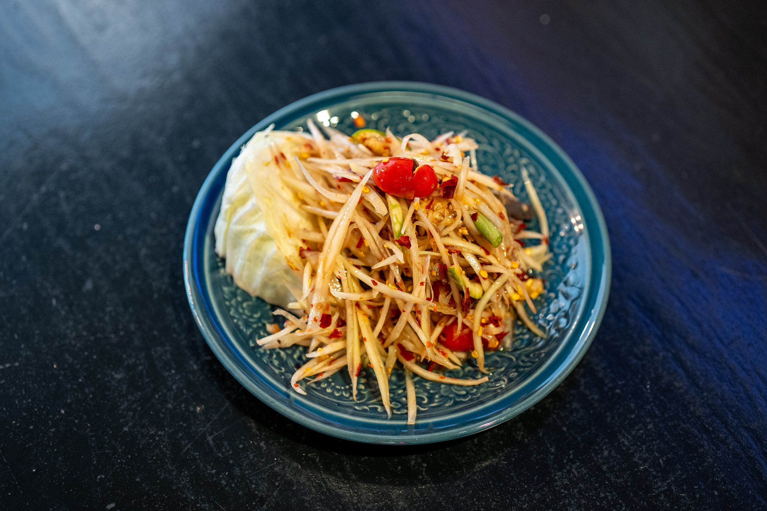 A blue bowl with papaya salad garnished with cherry tomatoes and chopped vegetables on a black surface.