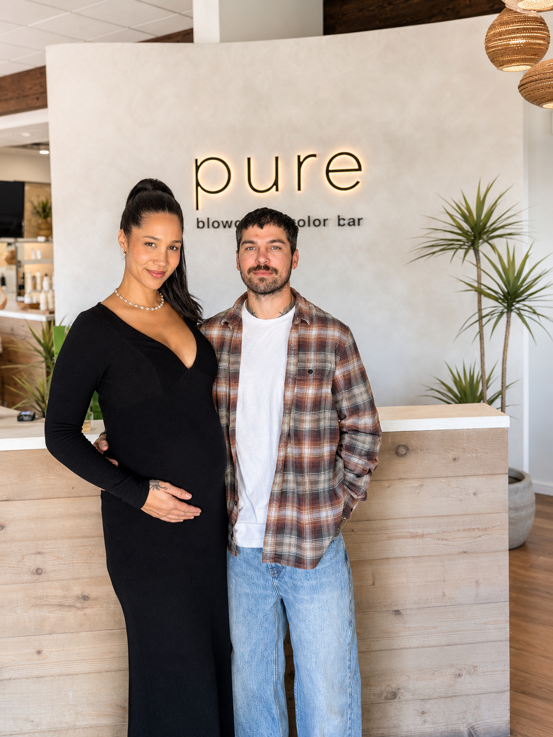 A pregnant woman and a man standing together in front of a reception counter at a salon called 'pure,' with the woman in a black dress and the man in a plaid shirt. The woman has her hand on her belly and both are smiling. The background features a modern salon interior with plants and soft lighting.