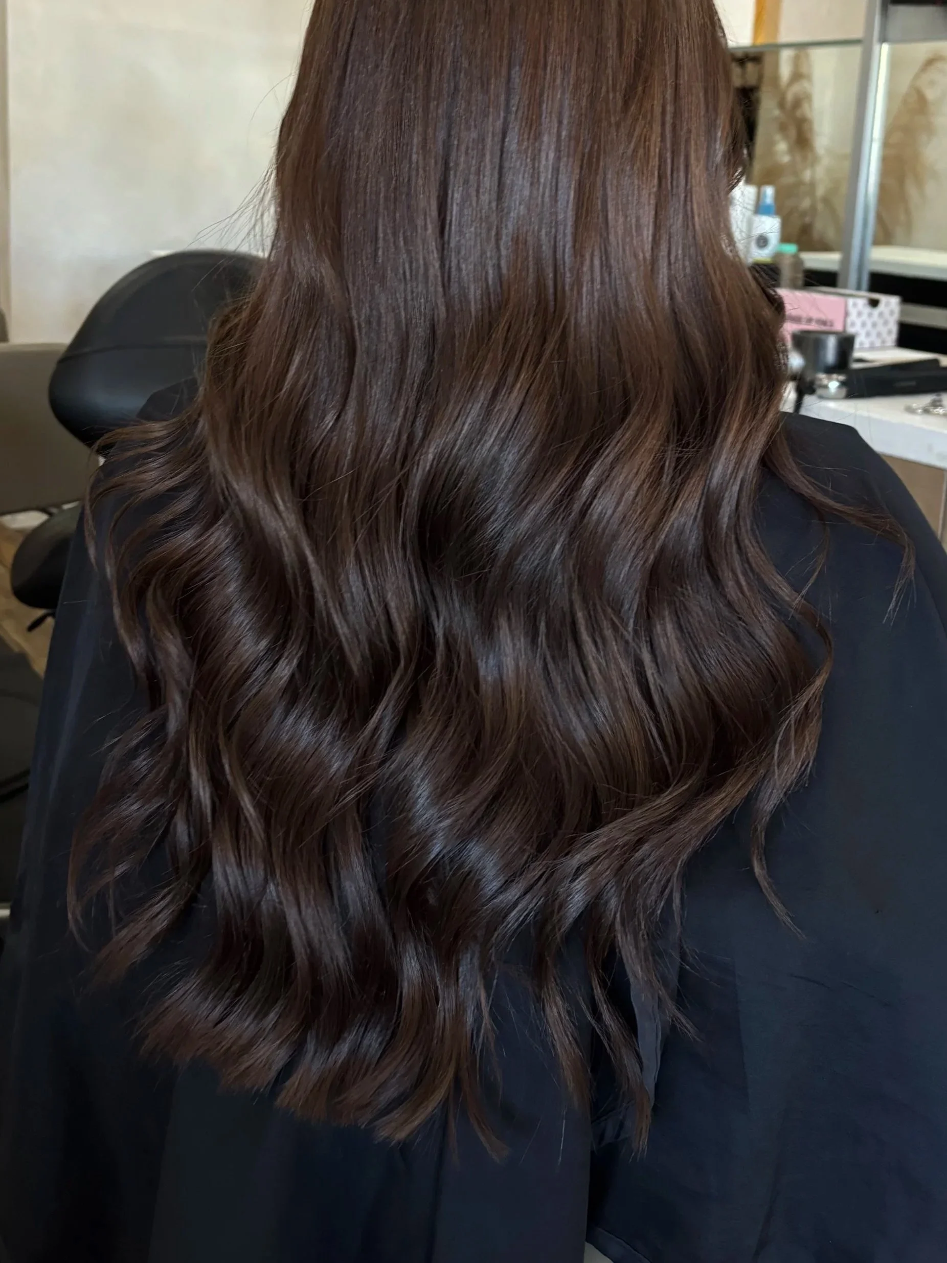 Back view of a person with long, shiny, brown wavy hair sitting in a salon chair.
