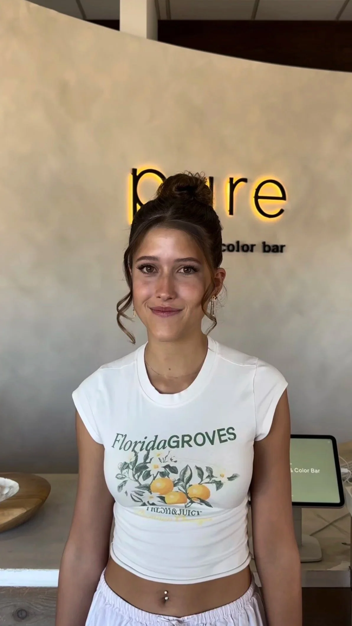 A young woman with brown hair styled in a bun, wearing a white sleeveless t-shirt with a citrus fruit and floral design, standing in front of a beige wall with yellow-lit dark text that reads 'pura' and smaller black text that reads 'color bar.'