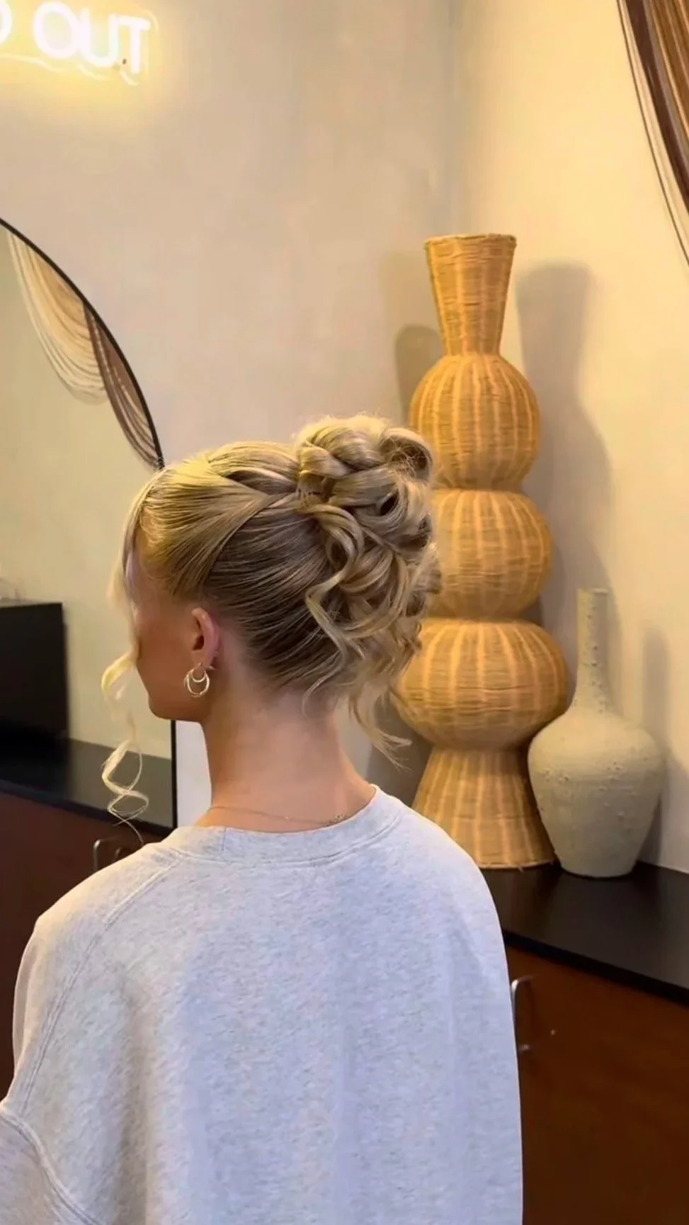 A woman with blonde hair styled in an intricate updo with curls and braids, wearing earrings and a gray shirt, stands in front of a tall, multi-tiered wicker vase in a room with neutral-colored walls and decor.