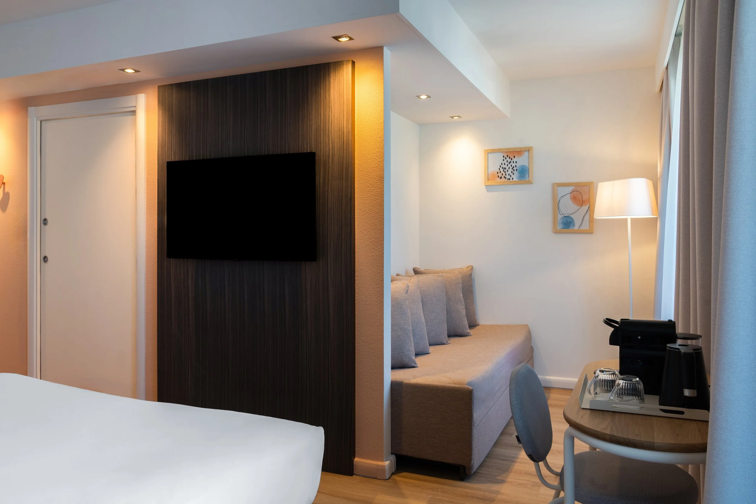 Coin salon dans une chambre d'hôtel avec canapé, télévision à écran plat, luminaires, œuvres d'art sur le mur, table avec machine à café et tasses, rideaux et lit partiellement visible.
